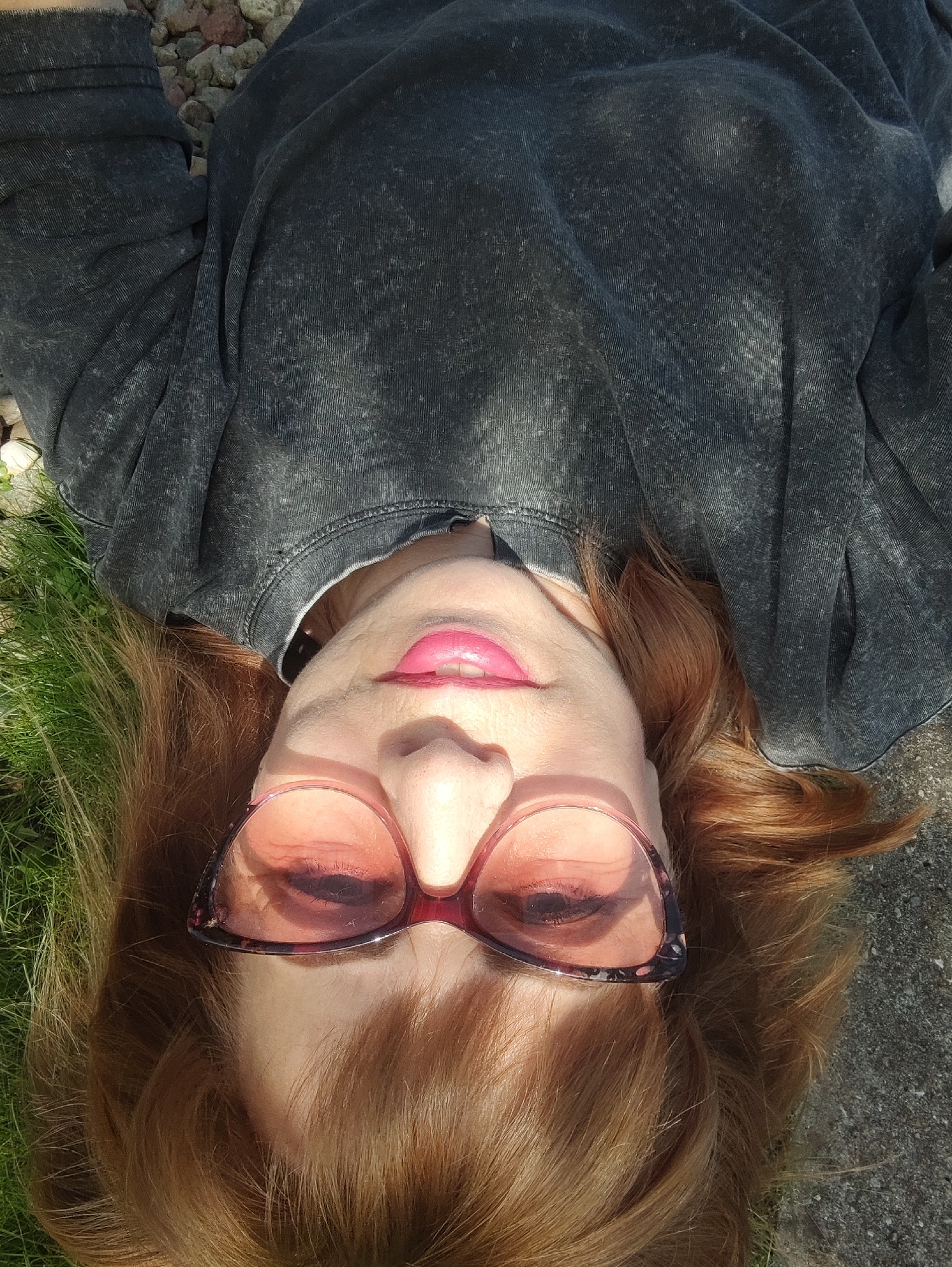 A woman with reddish-brown hair, wearing a dark gray sweatshirt and pink-tinted sunglasses, lies on the ground. She is outdoors with grass to her left and a gravelly surface behind her head. The sunlight casts shadows across her face and the sweatshirt. Her mouth is slightly open, revealing her teeth and what appears to be pink lipstick. The image is taken from a high-angle, looking down at the woman. The location is the city of Gvardeysk, Russia.