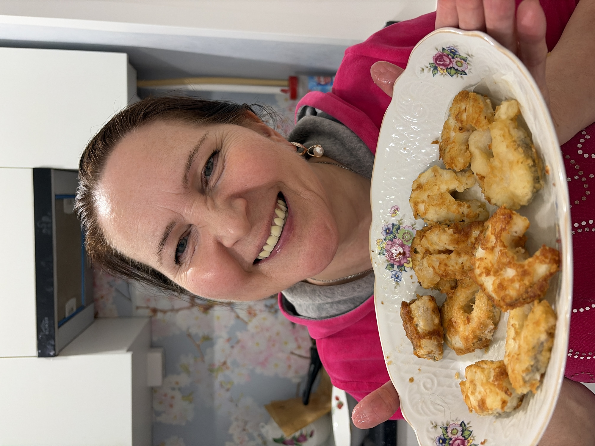 A woman with dark brown hair and a pearl earring smiles and holds a decorative oval plate of fried food. She wears a pink hoodie over a grey sweater. The background includes white cabinets and a wall with a floral wallpaper. The location in the city of Neftekamsk, Russia cannot be determined.