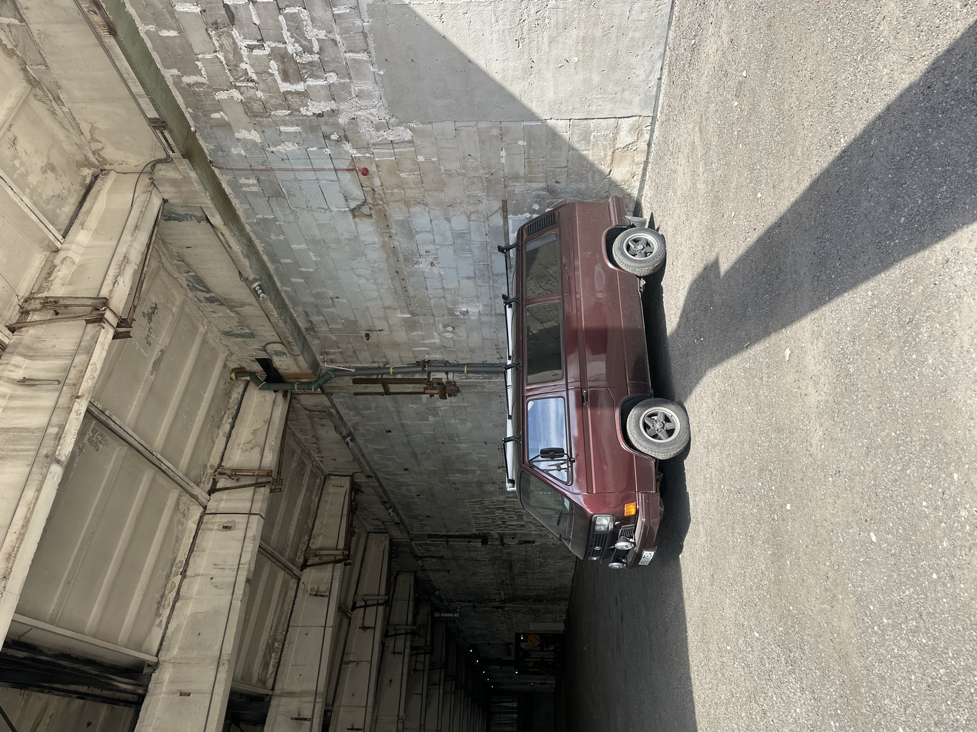 A maroon Volkswagen Vanagon, appearing to be a T3 generation model, is parked outdoors on a gravel surface. The van is positioned facing slightly to the left, with its front end visible. It has a roof rack installed. The environment is characterized by a large, industrial-looking structure with a concrete ceiling supported by beams and a wall constructed of large, rough-hewn concrete blocks. A long shadow cast by an unseen object stretches across the ground from the right side of the frame. The scene is well-lit by natural sunlight. The van's license plate is partially visible and reads "896 TEA". The architectural style of the building suggests it could be a former industrial facility or a bunker. Based on similar structures and the overall aesthetic, this location is likely the Linnahall building complex in Tallinn, Estonia, which is a former ice hall and concert venue known for its Brutalist architecture and underground areas.