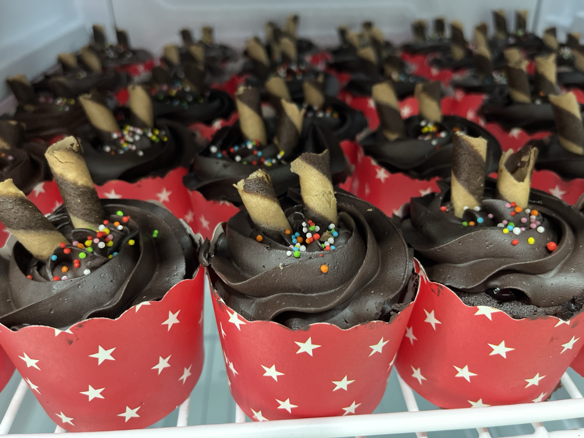 The image shows a close-up view of a shelf filled with chocolate cupcakes. The cupcakes are topped with dark chocolate frosting and decorated with colorful sprinkles and two striped wafer sticks. They are presented in red cupcake liners adorned with white stars. The cupcakes are arranged in rows, with some in focus and others blurred in the background, suggesting a commercial setting like a bakery or a market stall. The lighting appears to be from overhead. The environment suggests a cool, possibly refrigerated, display. The image does not provide any information about Lhokseumawe, Indonesia, or any specific location.