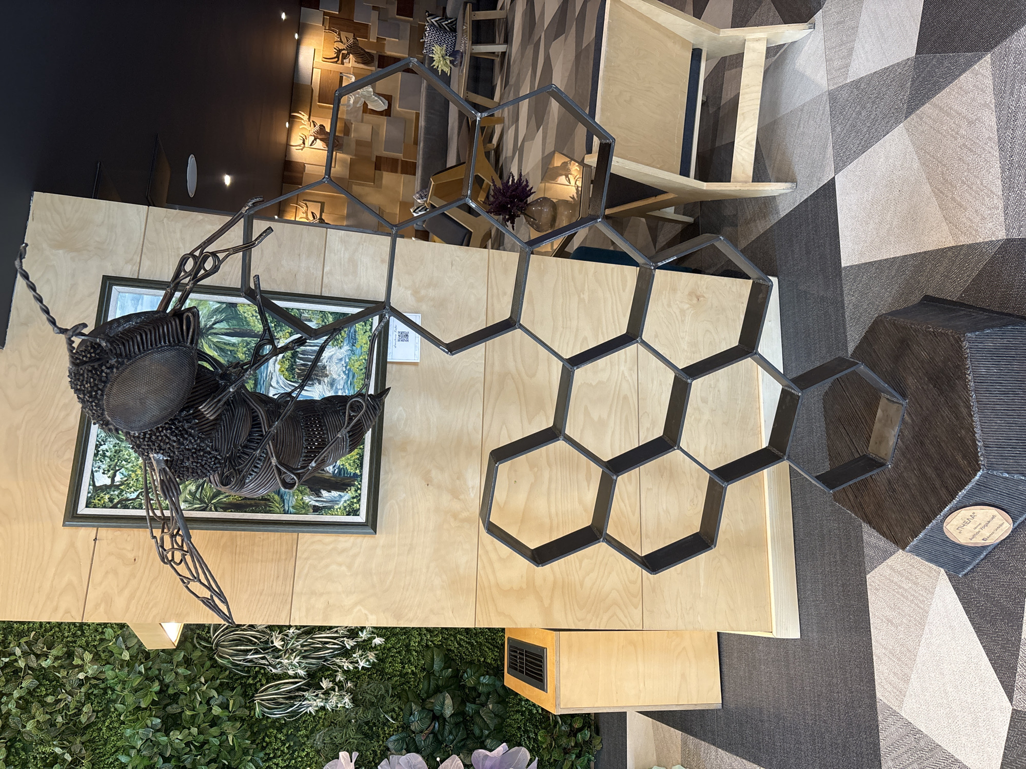 The image shows an indoor display featuring a metal sculpture of a bee positioned above a framed landscape painting, a decorative metal honeycomb structure, and a vertical garden panel. The honeycomb sculpture has a small placard attached to one of its cells. The environment appears to be a modern, interior space with light wood surfaces, textured carpet, and a black ceiling area. There is no indication of a specific city.