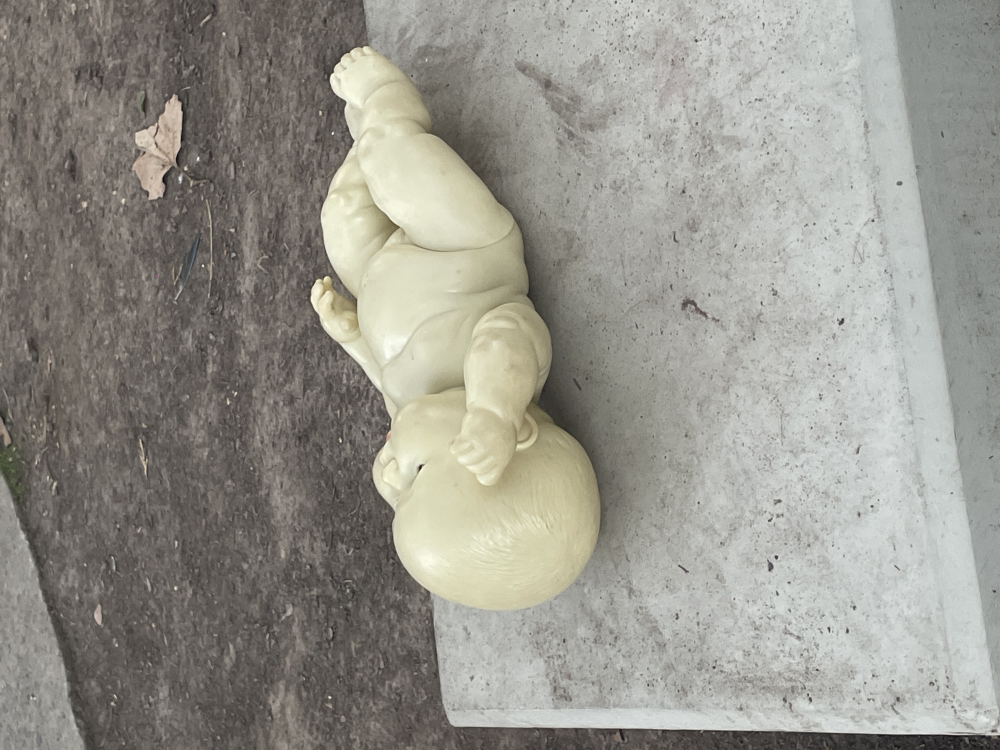 A light-colored, apparently plastic, baby doll lies on a concrete surface next to a patch of dirt in Olivos, Argentina. The doll is positioned upside down, with its head near the edge of the concrete and feet pointing away. A small, withered leaf rests on the dirt surface. The concrete surface appears stained.