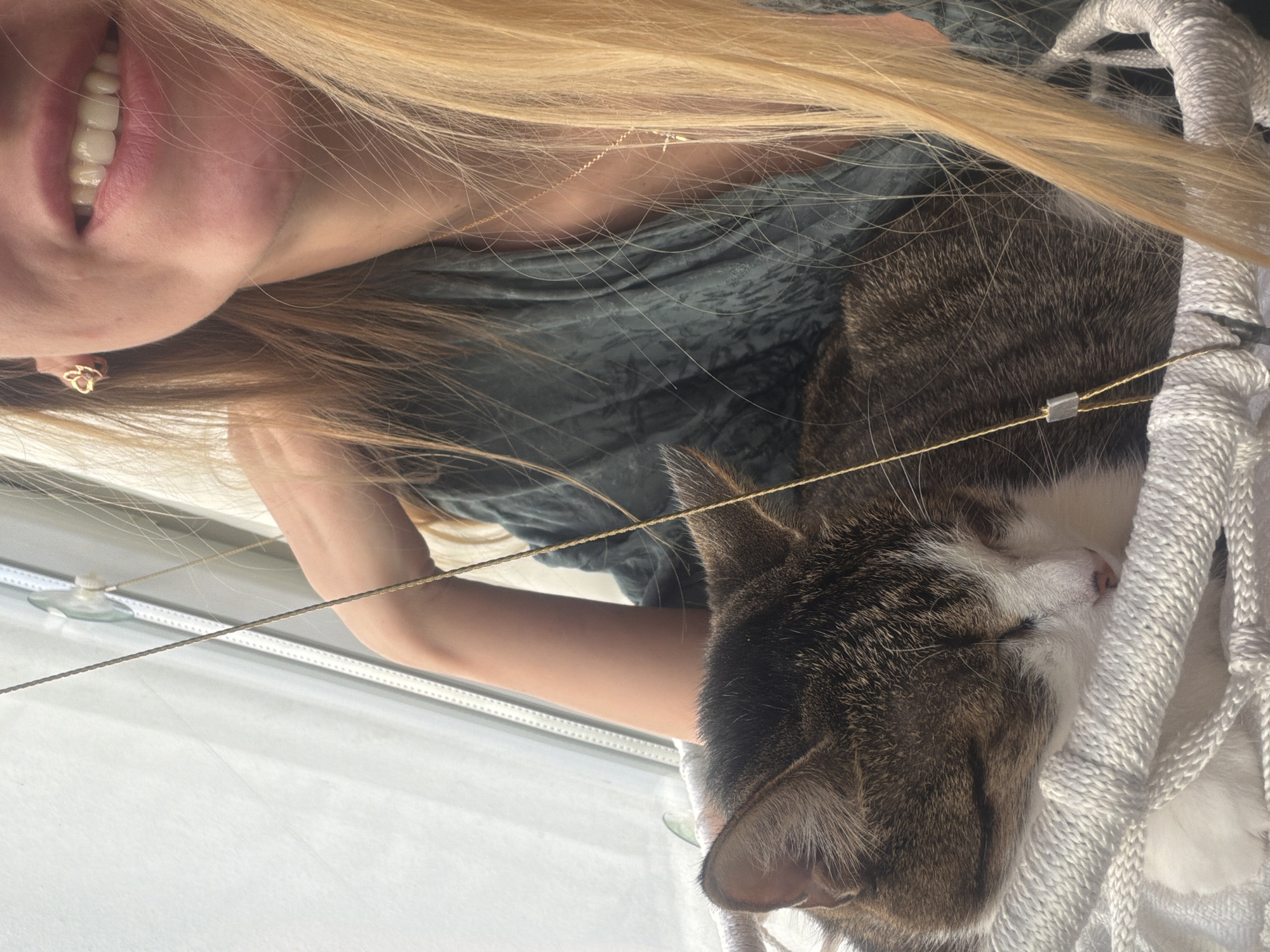 The image shows a close-up of a cat sleeping in a white knitted basket. A woman's face is visible in the upper right corner of the frame, with her mouth open in a smile and her blonde hair falling around her. The cat is a tabby with brown, black, and white markings. It is nestled in the soft, white material of the basket, appearing peaceful and comfortable. The woman is wearing a dark green top and a delicate gold necklace. The background is blurred, but suggests an indoor setting with a window. There is no indication of the city of Izmir, Turkey.