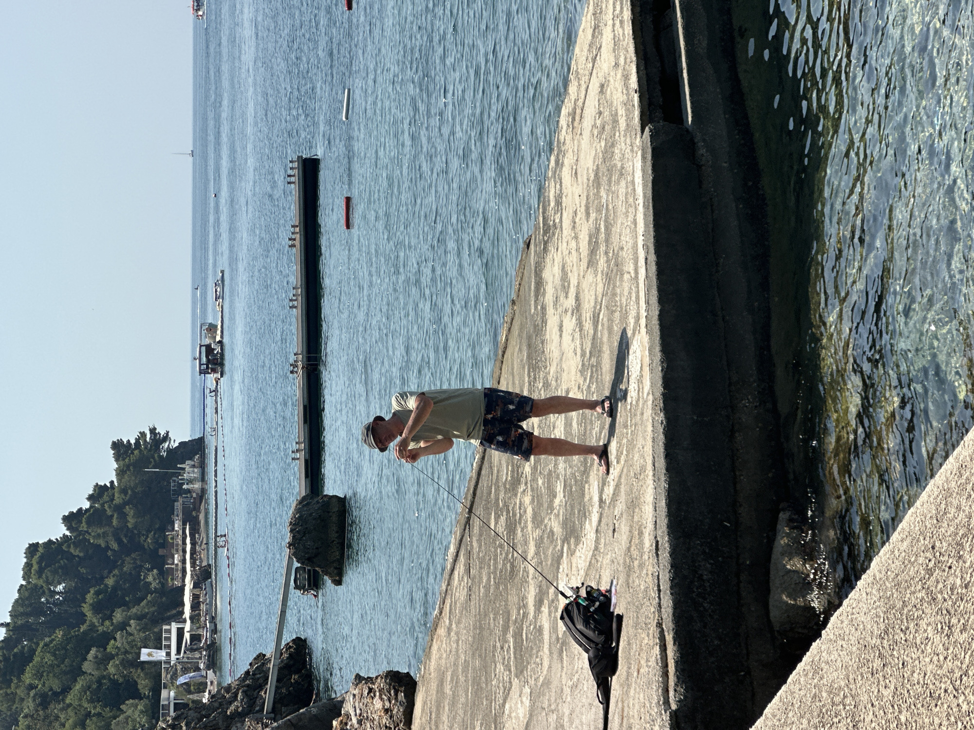 A man, wearing a light green t-shirt, dark patterned shorts, a wide-brimmed hat, and flip-flops, is standing on a concrete pier and appears to be fishing. He is holding a fishing rod and is looking down at it. A black backpack with fishing tackle is placed on the pier next to him. The scene takes place in Budva, Montenegro, with the Adriatic Sea in the background. Several boats and a distant sailboat are visible on the water, and a forested hillside with buildings and umbrellas lines the coast. The water is clear and blue, with some buoys marking a swimming area. The concrete pier is weathered and appears to be part of a waterfront area.