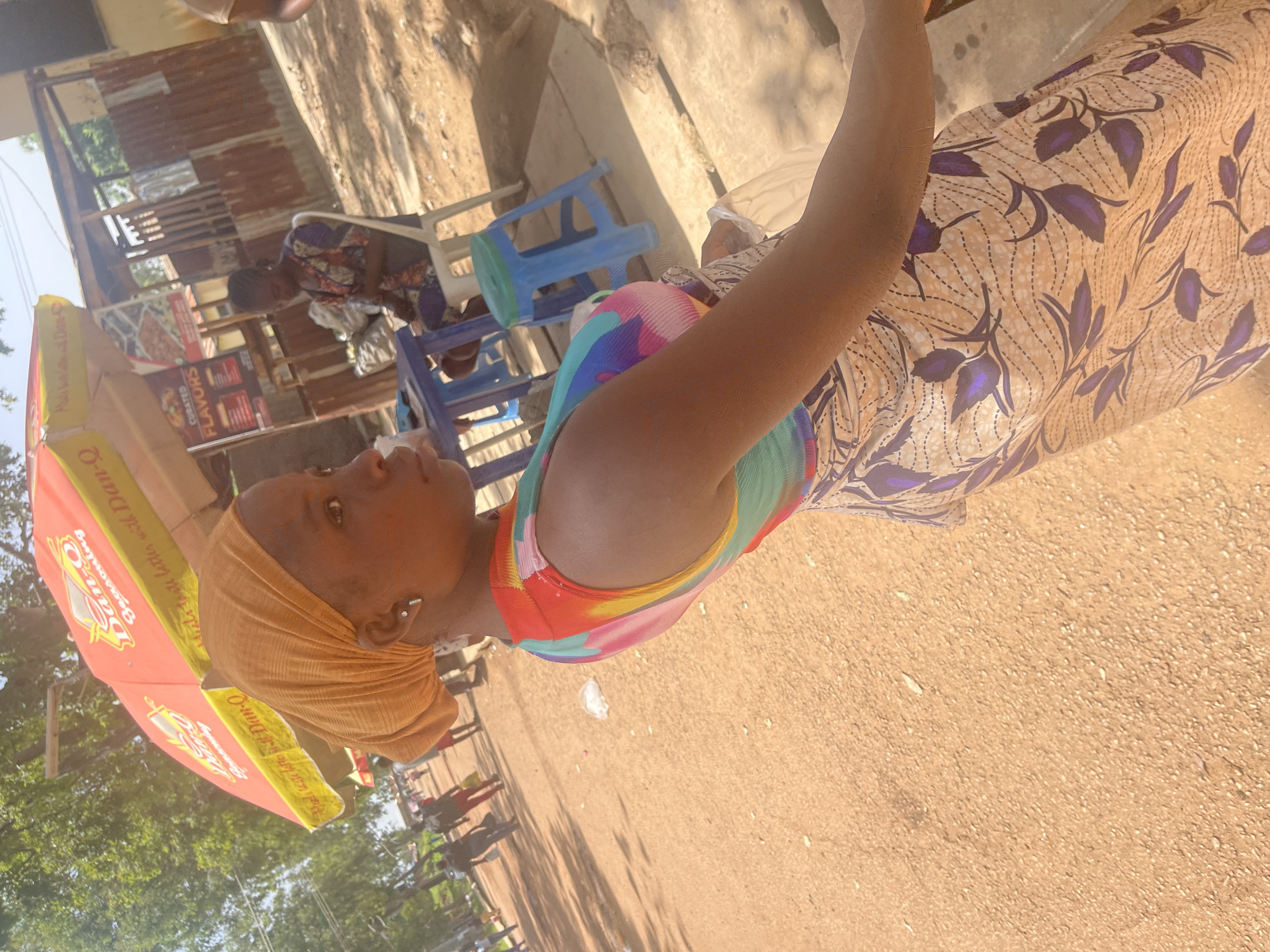 A woman wearing a patterned dress and an orange headscarf is the central subject of this outdoor image. She is positioned on the right side of the frame, with her body angled away from the viewer. Her left arm is raised, and she appears to be looking towards the left. She is wearing a colorful, sleeveless top underneath her dress, with vibrant patterns of pink, green, blue, and yellow. A small silver earring is visible in her left ear.
In the background, there are elements suggesting an informal outdoor market or roadside stall. A red and yellow umbrella is partially visible on the left, displaying text that includes "Mal, lagte letto ist Dan-Q," "Seasoning," and "FLAVORS." A corrugated iron structure is visible behind the woman, with what appear to be shelves holding various items. Blue plastic chairs are also present, some stacked. Other people are faintly visible in the background, some sitting and others standing.
The ground is a sandy or dirt surface, with scattered small white debris. The lighting suggests it is daytime, with shadows cast by trees and structures, indicating a sunny day. The overall scene conveys a casual, everyday moment in an outdoor setting, possibly a commercial area in Rafin Par, Nigeria, given the location context. The woman's expression is neutral.