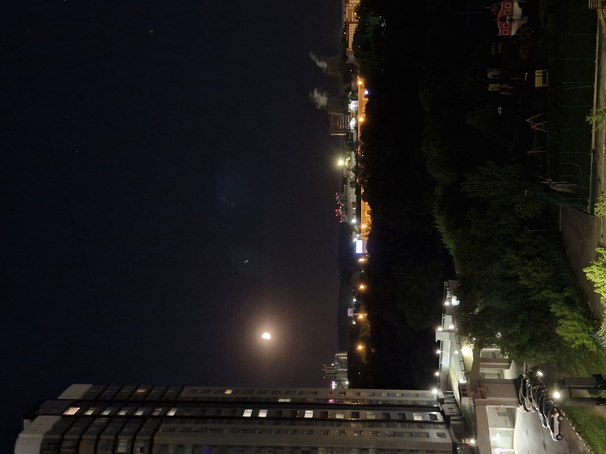 The image captures a night view in Yekaterinburg, Russia. The moon is in the sky, partially obscured. A large, multi-story apartment building is visible in the foreground. Across the street, there's a building with lit windows and a line of parked cars. Behind that, a dark area appears to be a green space, likely a park, with trees. Further in the distance, the city lights are visible, creating a horizon of lights. The sky appears dark, indicating it is nighttime.