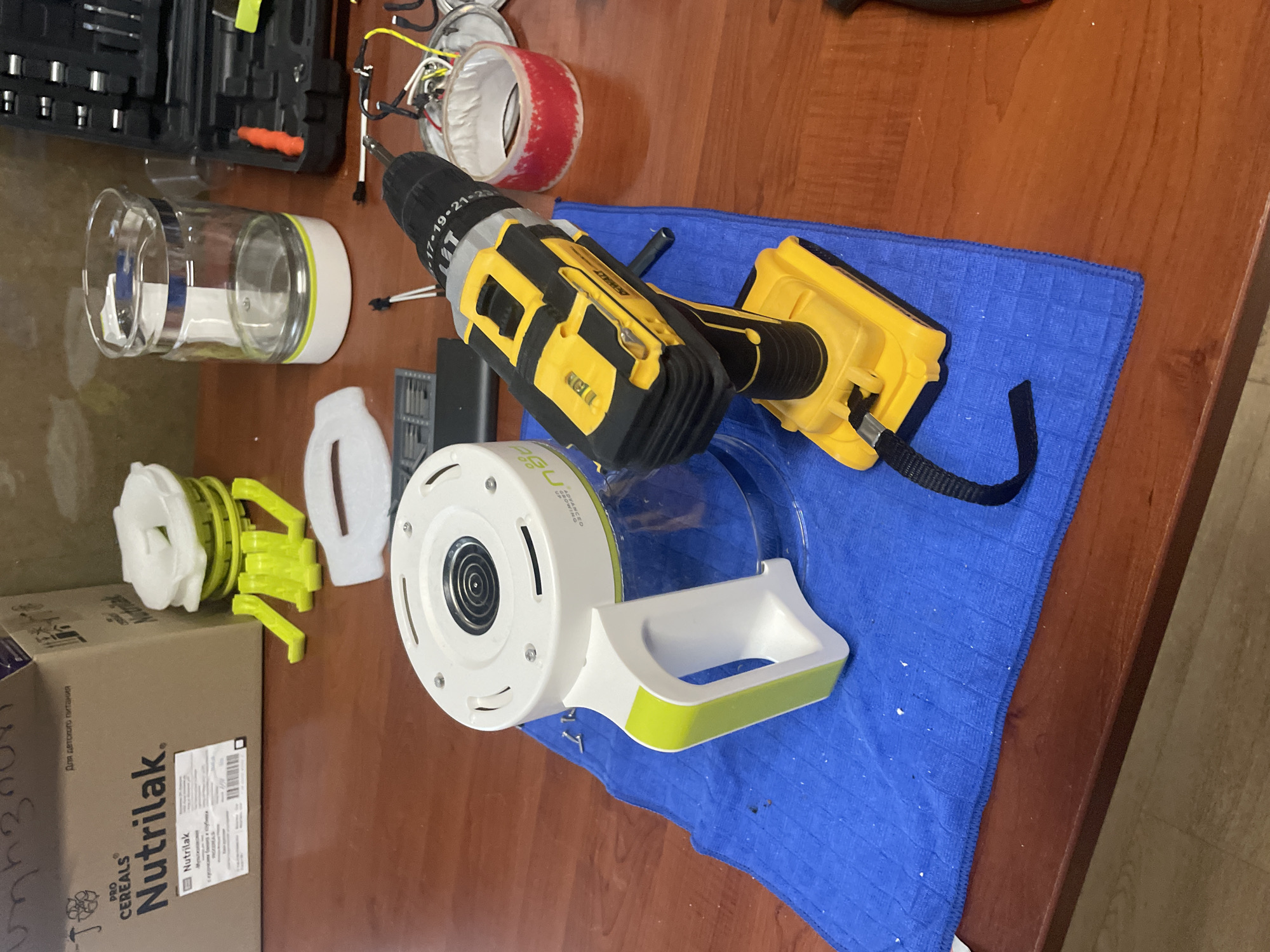 The image displays a wooden table with various objects. In the foreground, a yellow and black DeWalt cordless drill is positioned on a blue microfiber cloth. To the left of the drill, a white and green pitcher with a handle is also on the cloth. In the background, a cardboard box labeled "Nutrilak" and "PRO CEREALS" is visible. Next to the box are several white and lime green plastic parts, possibly components of the pitcher or another item. Further to the right, a clear glass container with a white base and a silver band sits on the table, alongside an open black toolbox containing various tools, including orange and red items. The lighting suggests an indoor setting, likely a home or workshop environment. There is no discernible indication of the specific location in Tbilisi, Georgia, beyond it being a flat surface indoors.
