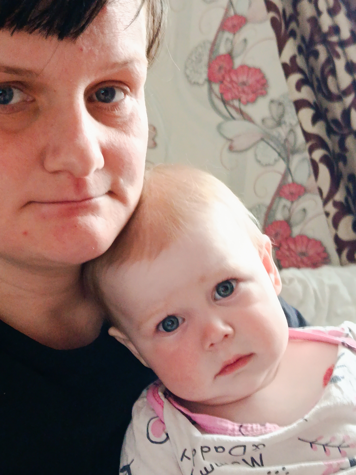 A close-up indoor portrait captures an adult woman holding a baby, both looking directly towards the camera. The woman, with short dark hair and blue eyes, occupies the left side of the frame, her face showing a neutral expression. Her right arm and shoulder are visible, clad in a dark top.
Resting her head against the woman's shoulder, the baby, with light reddish-blonde hair and blue eyes, is positioned on the right. The baby has fair skin and rosy cheeks and also maintains a neutral, somewhat curious expression. The baby is wearing a white top or onesie with a pink trim around the neck and various patterns, including parts of the words "Mommy" and "Daddy" clearly visible, along with letters like 'W' and 'C'.
The background features patterned wallpaper or a curtain with a floral design, depicting pink flowers and grey stems/leaves. To the far right, a dark brown patterned curtain is partially visible. The setting appears to be a cozy home environment. The lighting is soft and even, suggesting an indoor shot likely taken during the day or in a well-lit room. The image conveys a sense of closeness and familial warmth. This scene takes place in Фоки, Russia.