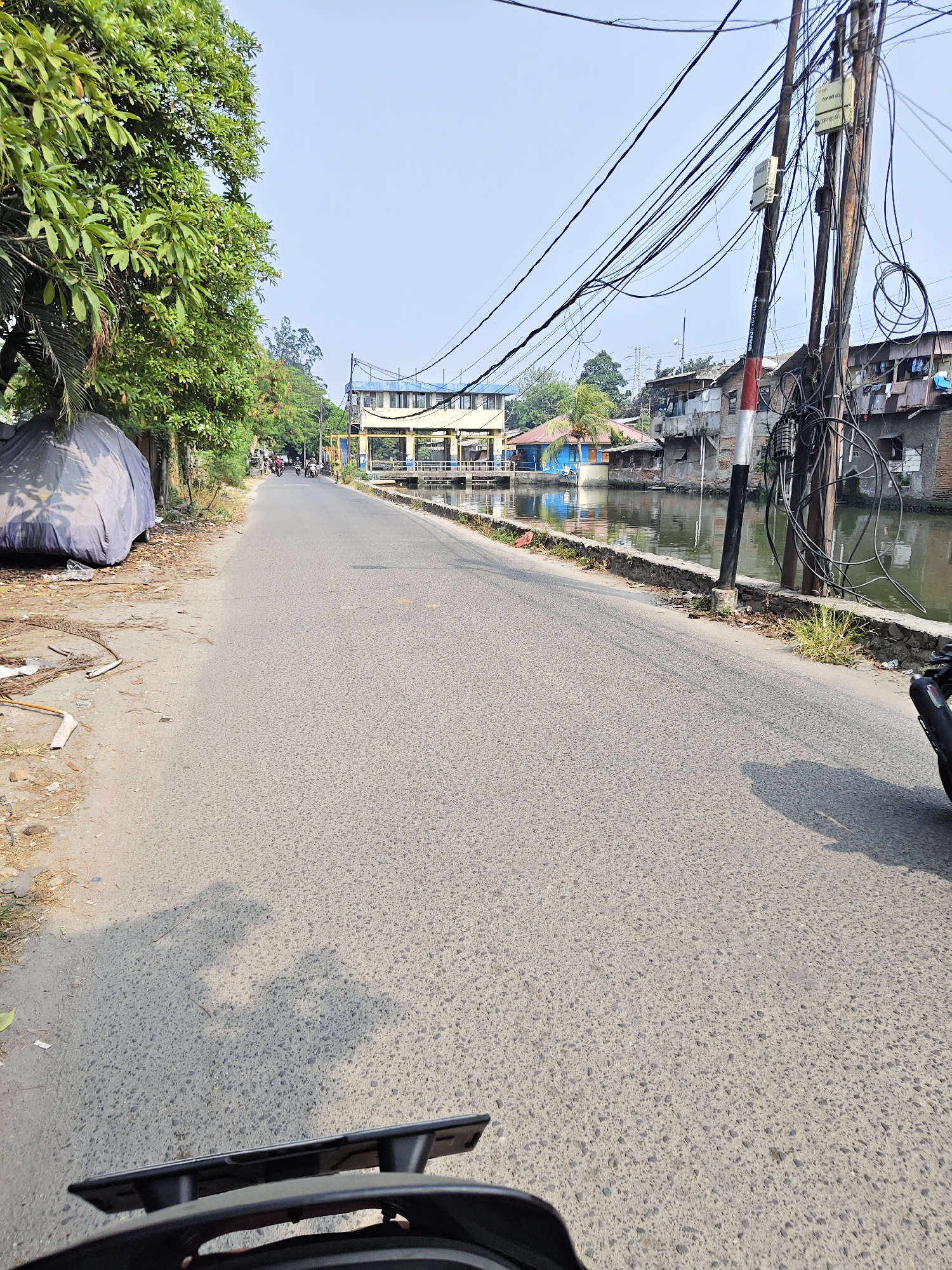 This is a photograph of a road in North Jakarta, Indonesia, likely near a canal or waterway. The perspective is from someone on a motorcycle, with the handlebars and a license plate visible in the foreground. The road is paved and appears to be in moderate condition, with visible texture and some debris on the side.
To the left of the road, there is a large tree with lush green leaves, and a blue tarpaulin covers an object, possibly a vehicle. Beyond the tree line, there are glimpses of other trees and vegetation.
On the right side of the road, a canal runs parallel to it. The water in the canal is murky. Along the canal bank, there are several buildings with corrugated metal roofs, characteristic of residential areas in Indonesia. A prominent feature on this side is a utility pole heavily laden with a dense network of electrical and communication wires. The pole itself has a red and white striped band near its base.
In the distance, a yellow pedestrian bridge with a blue roof spans across the canal. There are a few motorcycles and people on the road, indicating ongoing activity. The sky is clear and bright, suggesting a sunny day. The overall scene depicts a somewhat cluttered but functional urban environment, typical of many areas in Jakarta.