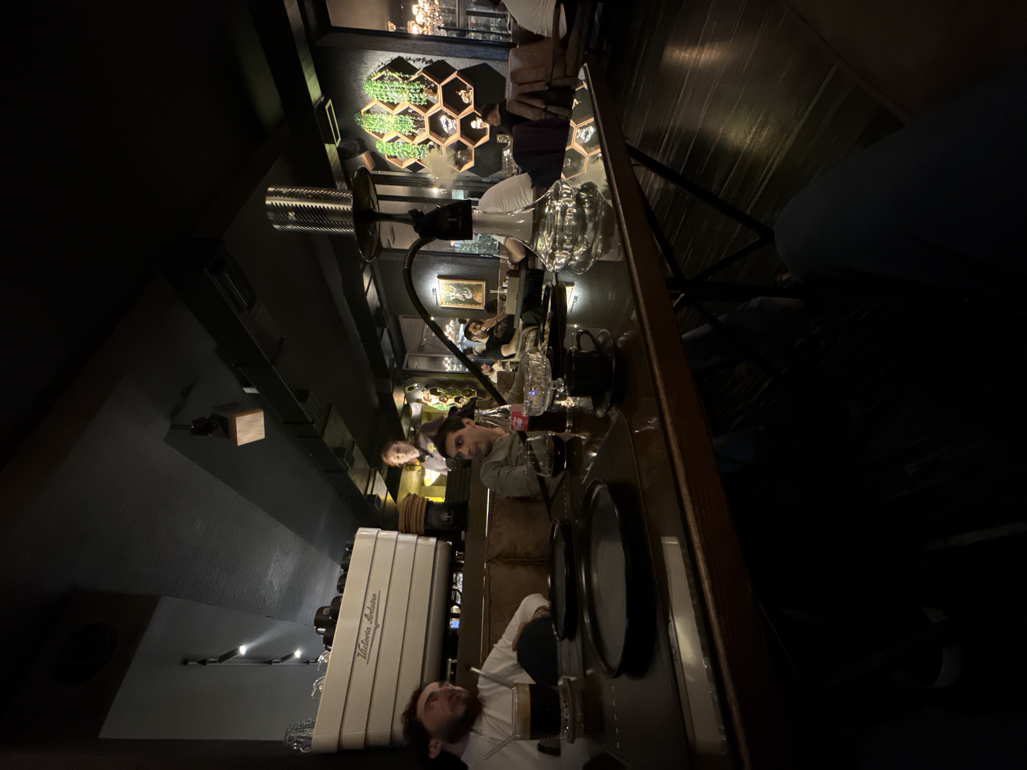 Here is a matter-of-fact description of the image:
The image shows the interior of a dimly lit establishment, potentially a cafe or lounge. Several people are present at a table, appearing to be engaged in conversation and possibly enjoying refreshments. Visible on the table are glasses, plates, a Coca-Cola bottle, and what appears to be a hookah. One individual is wearing a white shirt and dark cap. The surrounding décor includes a green wall feature with plants in geometric displays, a coffee machine with the brand name "Victoria Arduino" on its side, and overhead lighting. The scene suggests a relaxed, social atmosphere.