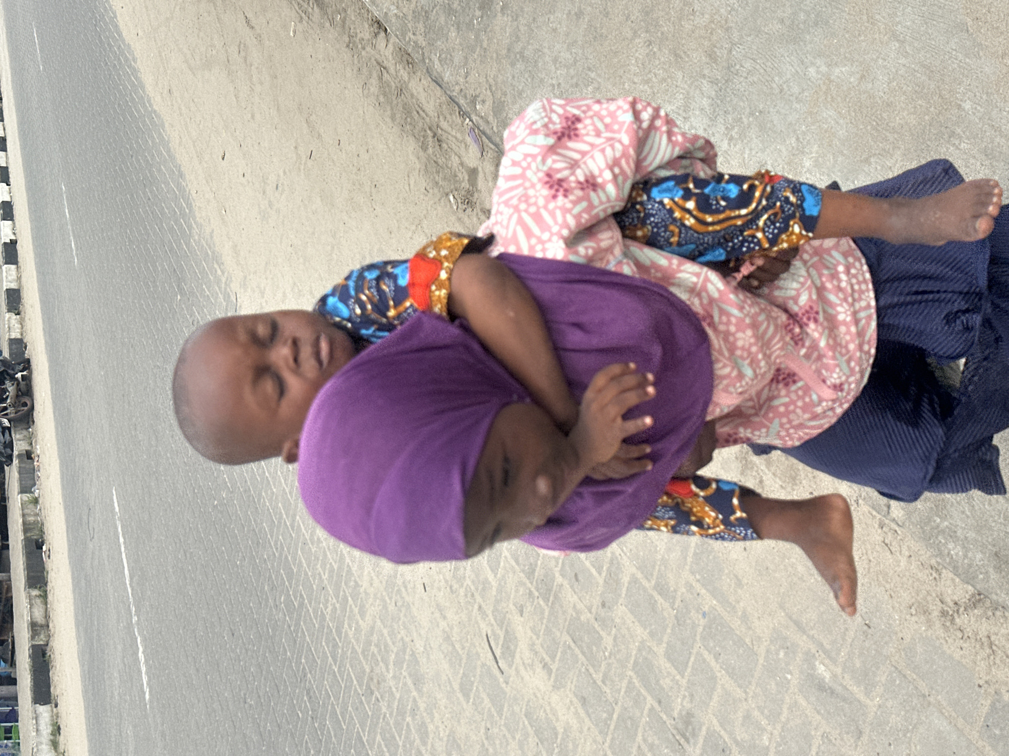 An outdoor, eye-level, medium shot captures two young children from the chest up on a paved sidewalk adjacent to a road. The child in the foreground, wearing a dark blue skirt and a pink and white patterned top, carries the other child on their back. The child being carried is wearing a purple headscarf and a patterned blue and orange shirt. Their eyes are closed, and they are resting their head on the carrier's shoulder. The carrier's face is partially obscured, looking down, with their arms wrapped around the child on their back. The road in the background has a white line marking and is bordered by a concrete curb with a gray, brick-patterned walkway. In the far background, a few indistinct objects and what appears to be a motorcycle are visible. The overall lighting suggests it is daytime.