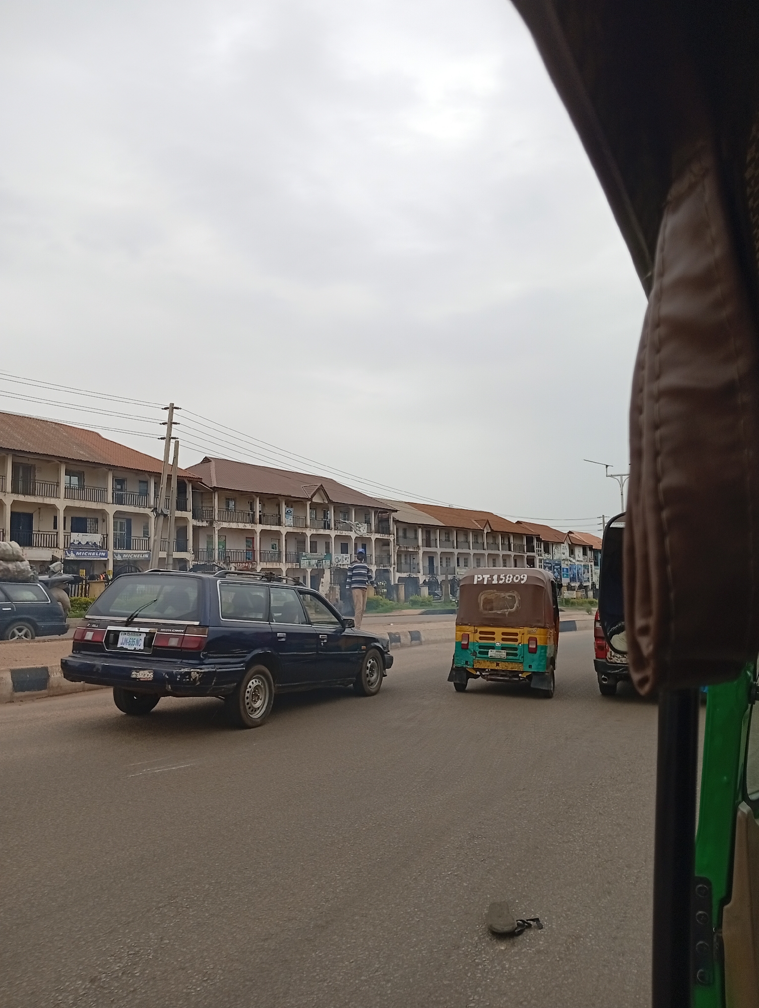 In Gura Top, Nigeria, a street scene unfolds under an overcast sky. A dark blue station wagon, identified by license plate LJN-635 NC, travels down a paved road. To its right, a three-wheeled auto-rickshaw, displaying license plate PT-15809, follows. Further ahead, another auto-rickshaw is visible. The road is bordered by a row of multi-story buildings with balconies and brown roofs. On the left side of the road, a dark car is parked, carrying a load of sacks. A lone man stands near the buildings, conversing with a person who is partially obscured. The perspective suggests the photograph was taken from another vehicle.