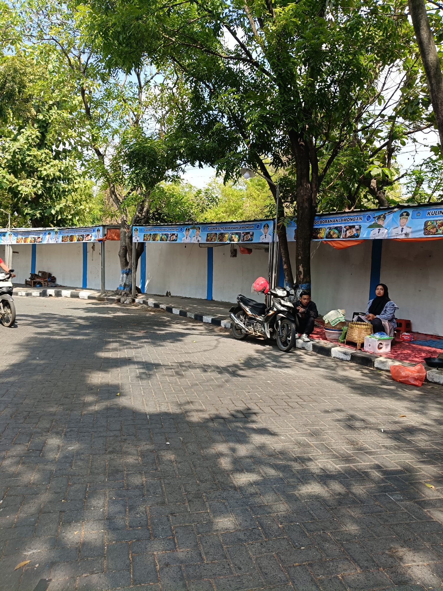 This image depicts a street scene in Lamongan, Indonesia, likely near a culinary area or market. The foreground is a paved walkway with a herringbone pattern, shaded by trees. A motorcycle is parked on the walkway, to the left of two individuals sitting on the ground.
In the middle ground, a white wall with blue trim serves as a backdrop. Above this wall, a banner displaying images of food and official portraits is visible, suggesting a food stall area. Two people are seated on red mats in front of the wall: a man wearing a black shirt and black pants, and a woman wearing a blue hijab and a blue patterned top. They are surrounded by baskets and bags, and a red helmet rests on the motorcycle.
Further down the wall, empty stalls are visible, with some discarded wooden chairs and other items in front of them. The overall environment appears to be outdoors, under a bright sky with sunlight casting shadows. The presence of the banner with "KULINER SEGO BORANAN LAMONGAN" indicates the specific location is associated with the regional specialty of Sego Boranan, a rice dish from Lamongan.