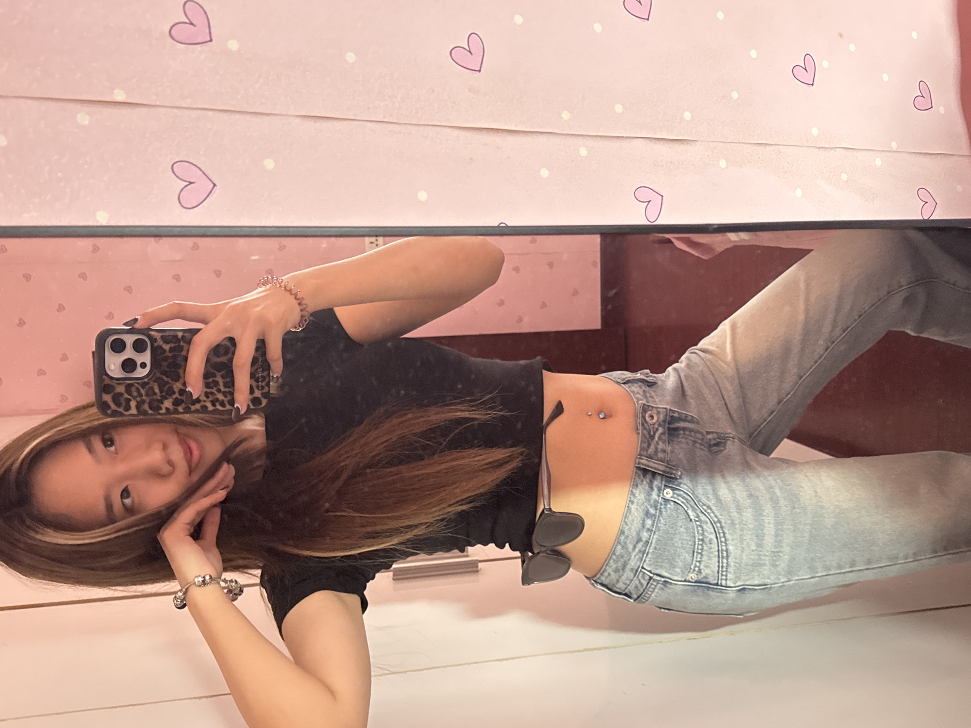 A person, presumably female, is reflected in a mirror while taking a selfie. The person has medium-length brown hair with blonde highlights and is wearing a short-sleeved black top and light blue jeans with a navel piercing. Sunglasses hang from the waistband of their jeans. The person holds a phone with a leopard-print case. The background behind the mirror features a pink surface decorated with white polka dots and purple hearts. It's impossible to determine the location in the city of Shapingba District, China, from the provided image.