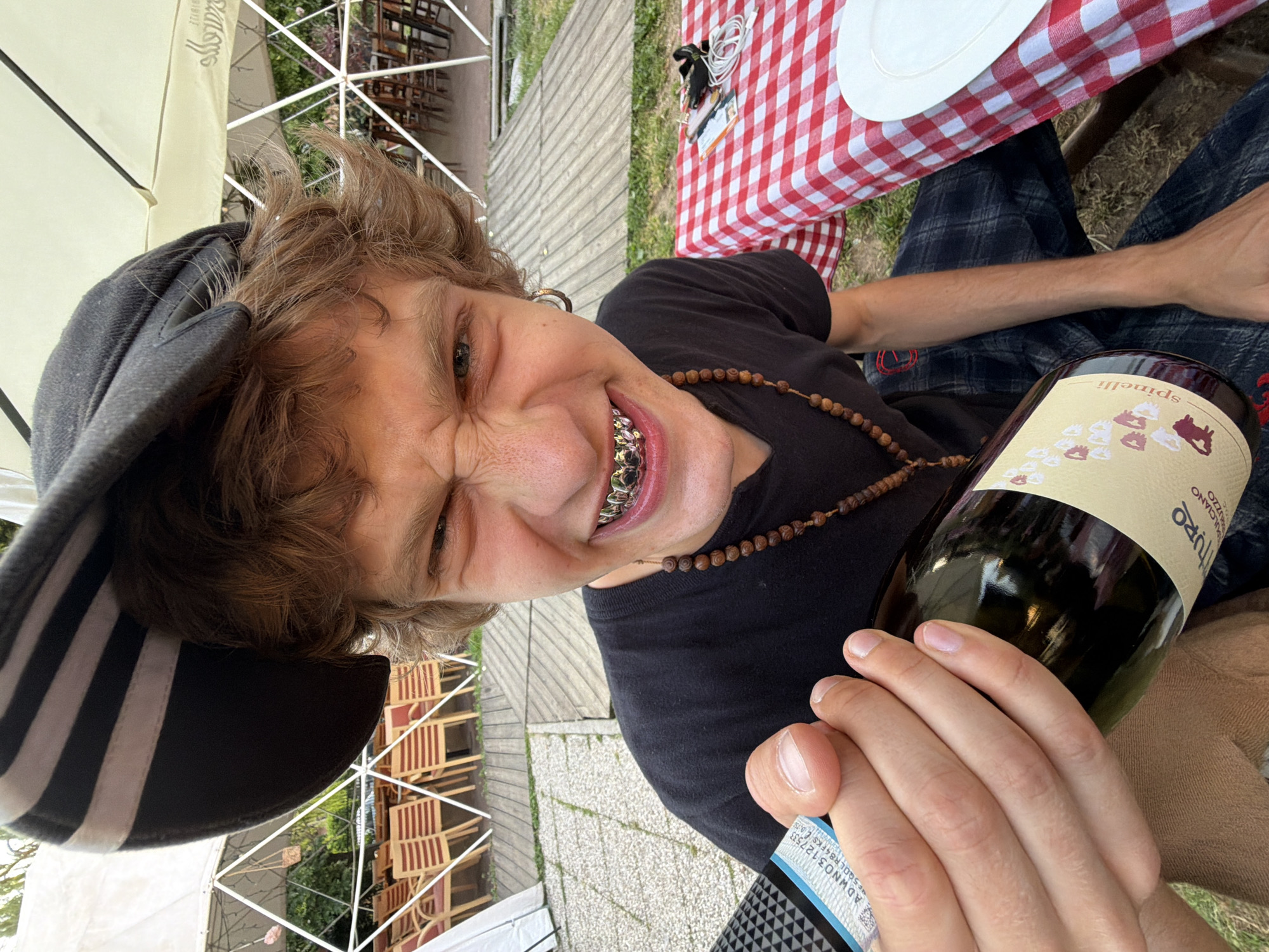 A dynamically angled, close-up shot captures a young individual seated outdoors, holding a bottle of wine. The person, with medium-length brown hair, wears a dark cap with white stripes on the brim's underside and a black T-shirt with a wooden beaded necklace. They look directly at the camera with an exaggerated, playful grin, showcasing metallic grillz on their upper teeth.
The right hand firmly holds a dark glass wine bottle. The label partially reveals "Spinelli" on the neck (inverted) and "SPINELLI" along with "VURVEGIO" (likely Verveglio) on the main label. A blue sticker with "ADWN03-12753" is also visible on the bottle.
The setting is an outdoor or semi-outdoor casual dining area, possibly a patio or garden in Milan, Italy. A red and white checkered tablecloth covers a table, where a white plate and some cables are visible. The ground is paved with a light-colored cobblestone-like pattern. In the background, stacked wooden chairs and trees suggest a restaurant or cafe. A large, light-colored outdoor umbrella, with barely visible text "Silv...", provides shade. The natural lighting indicates it is daytime, and the overall mood is informal and expressive, highlighted by the subject's humorous pose.