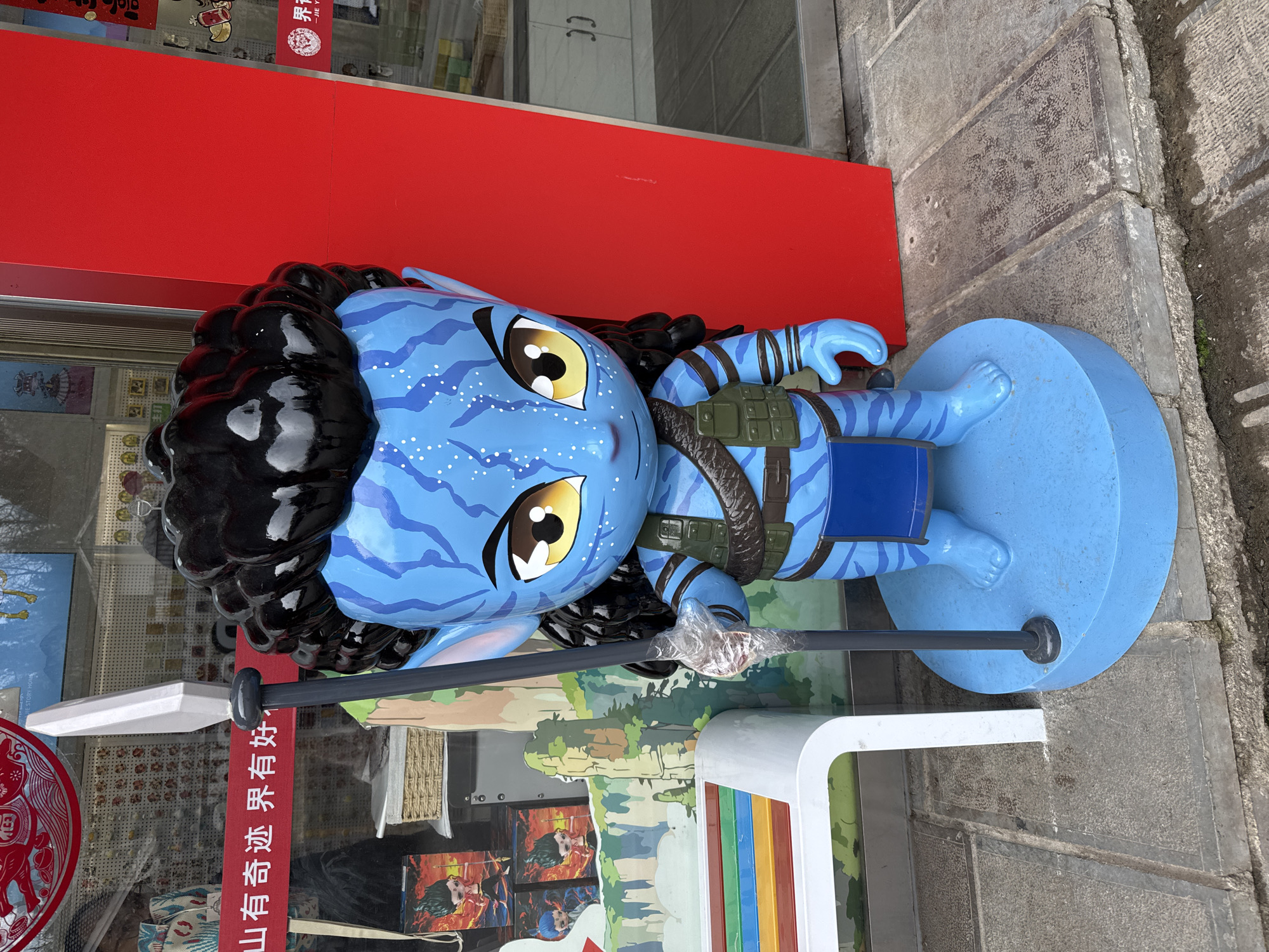 An outdoor, eye-level shot in Wulingyuan, China, displays a stylized, blue-skinned humanoid statue resembling a Na'vi character from the *Avatar* film series. The statue, approximately life-size, stands on a circular blue base on a grey stone sidewalk. It features a large head with black, wavy hair, prominent yellow eyes, and a patterned blue face with white dots. The body is clad in a dark green vest with pouches, a brown belt, and a blue loincloth. Its right hand, wrapped in clear plastic, grips a tall, grey spear with a white, pointed tip.
To the immediate left of the statue, a white bench with multi-colored horizontal slats (red, orange, yellow, green, blue) is partially visible. Behind the bench and the statue, a storefront window displays various merchandise and promotional graphics. A red banner with white Chinese characters reads "山有奇迹 界有好" (Mountains have miracles, the world is good). Further within the window, illustrations of floating mountains and an animated character are visible, suggesting a souvenir or tourist shop.
To the right of the statue, a bright red vertical panel or wall section is present. Beyond this red panel, another storefront window is visible, showcasing additional items and a partial red and white circular logo. The ground consists of uneven grey concrete or stone slabs, with visible cracks and minor debris.