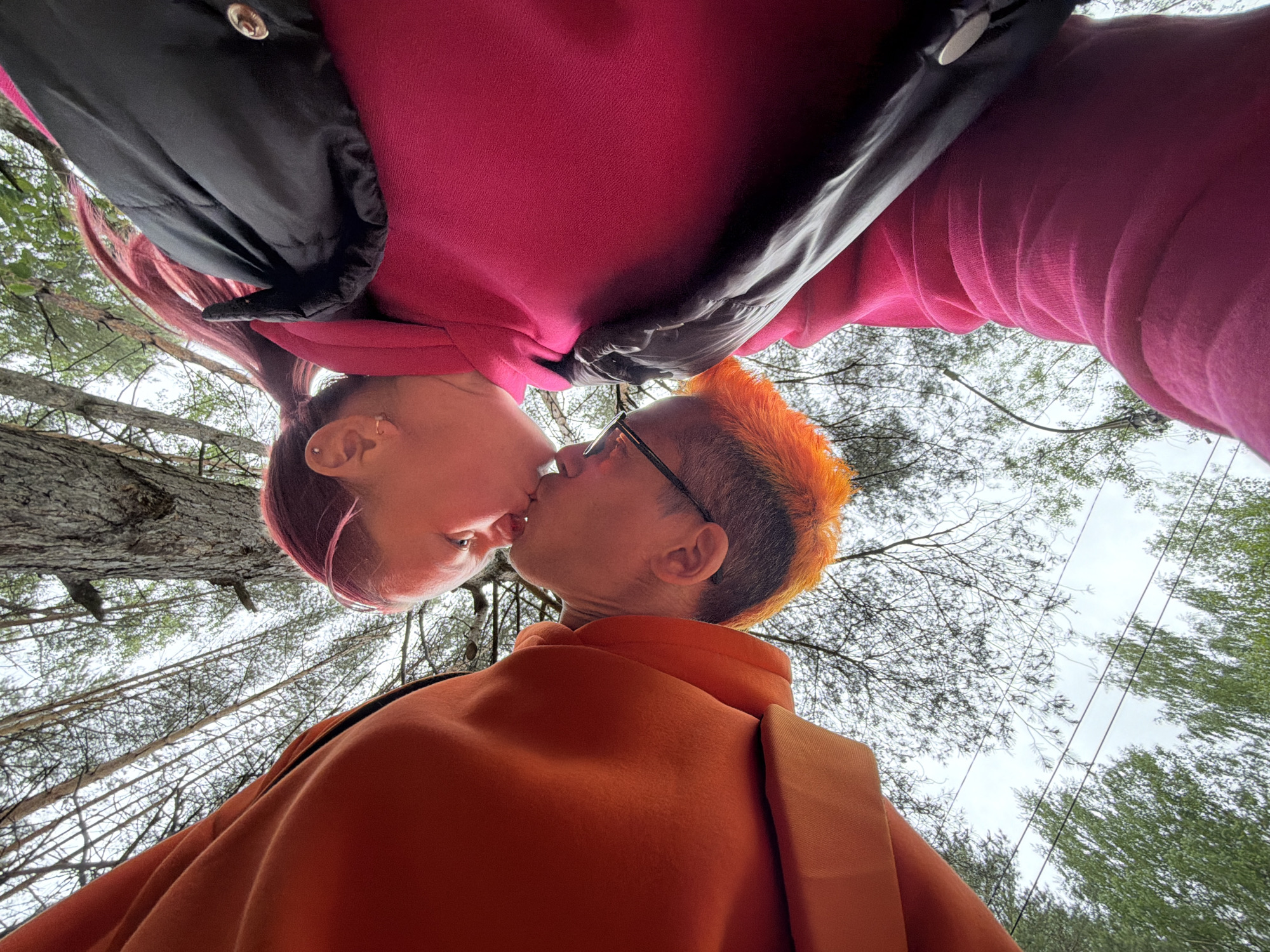 A low-angle shot depicts two individuals kissing outdoors. The person on the bottom wears an orange jacket and eyeglasses, and has orange hair. The person on top wears a pink jacket and a black vest, with reddish hair. The background consists of trees and sky. The location is Chaikovsky, Russia.