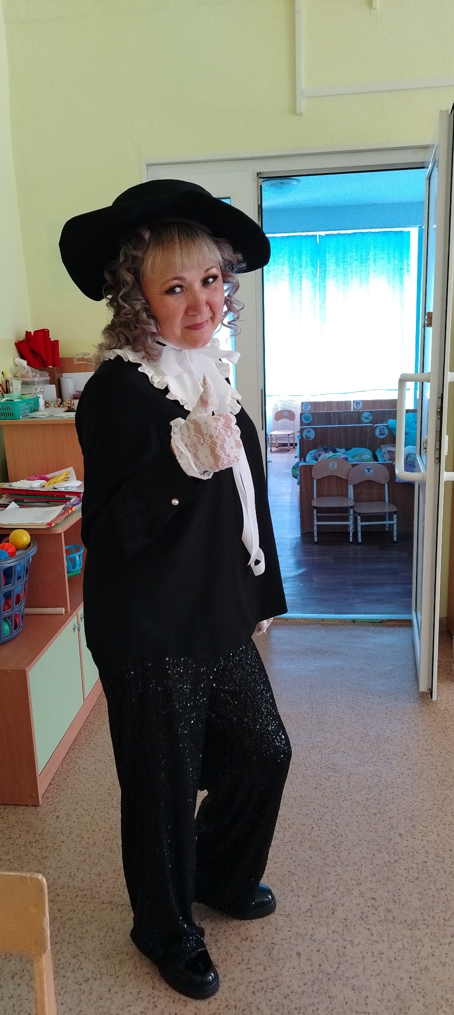 A full-length shot depicts a woman dressed in a theatrical costume. The woman is wearing a wide-brimmed black hat, a black blazer with a white, ruffled collar and a white lace embellishment on her hand. She's also wearing sequined black trousers and shiny black shoes. The woman is standing near an open doorway in a building that appears to be a classroom with children's furniture and supplies. Based on the analysis of other images, the location is likely a kindergarten in Chaikovsky, Russia. The woman is looking towards the camera with a subtle smile.