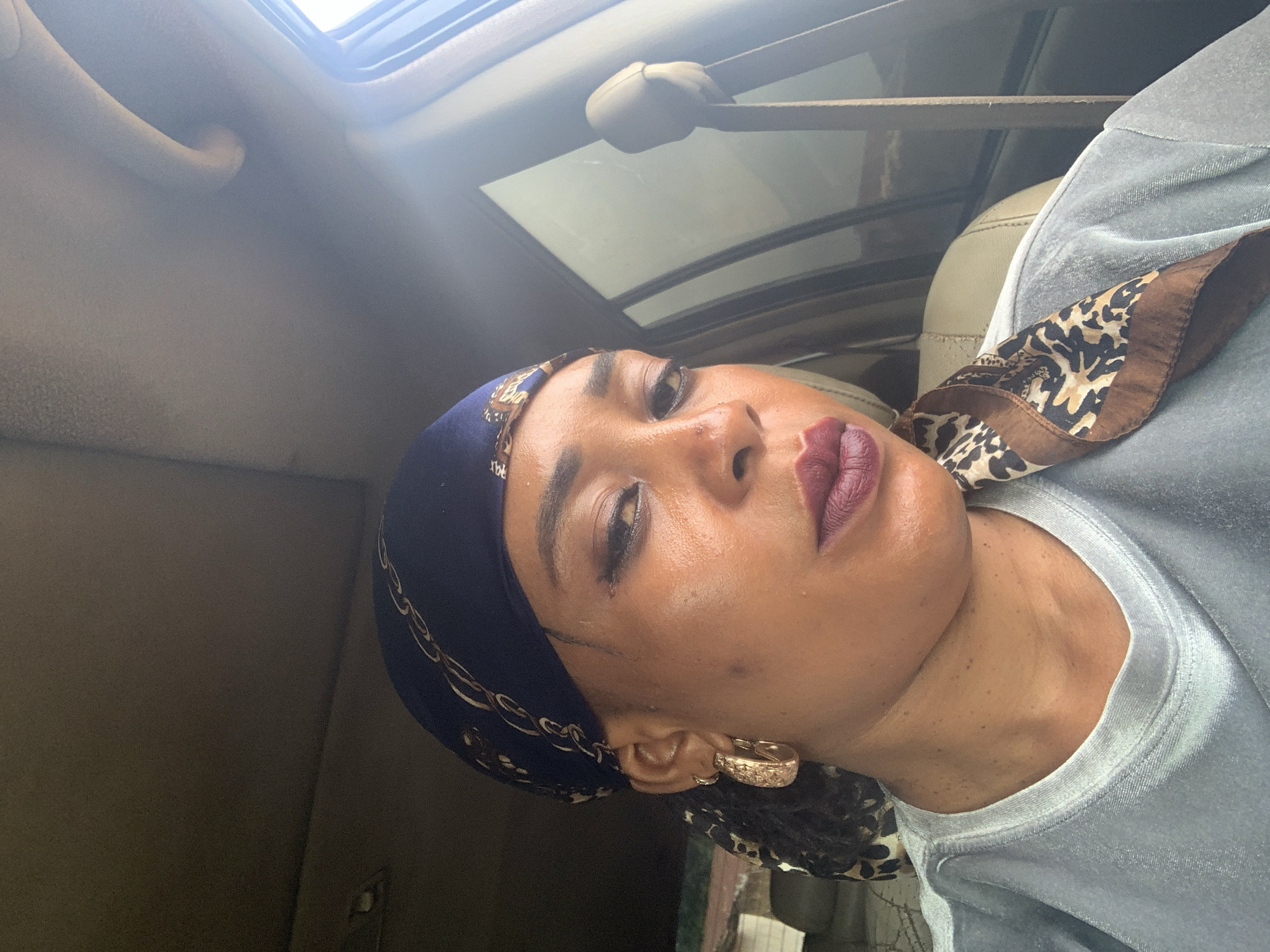 A person is in a vehicle with a sunroof. The person has on a dark blue head scarf with a gold chain pattern. The person is wearing a gray shirt with a leopard-print scarf attached. The person is wearing dark red lipstick and gold earrings. The environment shows parts of the car interior, including the sunroof, handle, and seat. There is no information in the image to determine the location in Sabon Tasha, Nigeria.