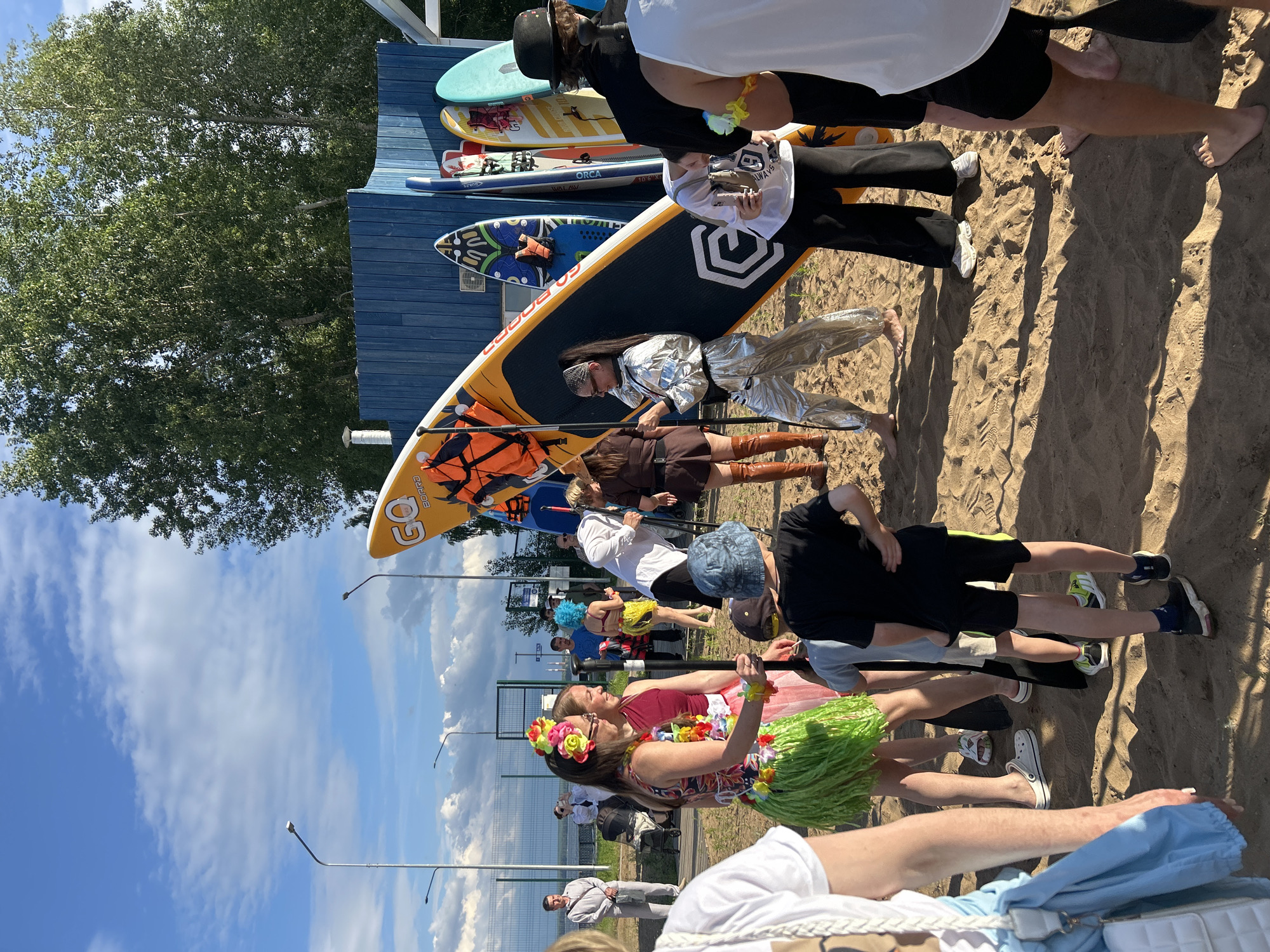 This is a horizontal, eye-level, full shot of a sandy area with several people and paddleboards. A blue building is in the background, with trees and a partly cloudy sky above. A group of people are gathered near the building and several paddleboards are propped up against it. In the foreground, a young person in a black t-shirt and shorts stands on the sand, facing away from the camera. A woman in a green grass skirt and a colorful top, adorned with flowers and leis, stands to the left of the frame, facing right and smiling. She is holding a paddleboard. Behind her, a person in a red top and white skirt is also smiling. To the right, a person in a silver metallic suit is posing for a photo. Other individuals in various attire are scattered throughout the scene, some interacting with each other, others standing near the paddleboards. The lighting suggests it is daytime, likely late afternoon due to the long shadows cast on the sand. The presence of paddleboards indicates a potential watersport or beach-related event. Based on the context from a broader dataset, this scene is likely located at a beach or watersport club in the city of Chaikovsky, Russia, possibly near the Kama River reservoir, which is a popular recreational area in the region.