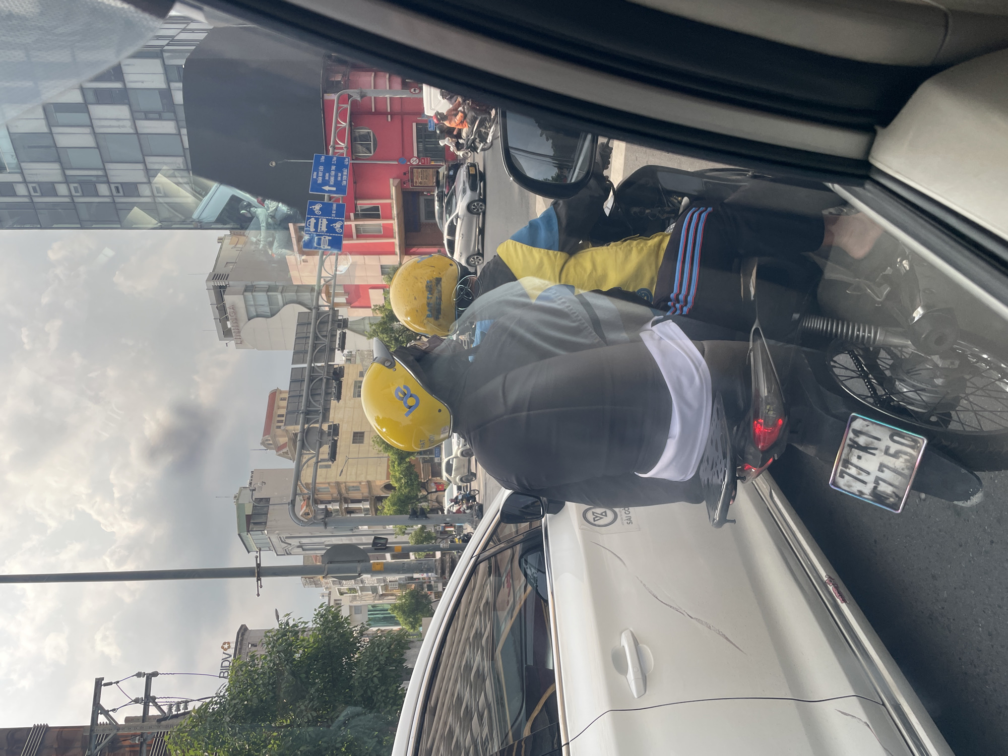 The image depicts a street scene in Thủ Đức, Vietnam, likely during the daytime under cloudy skies. In the foreground, viewed from inside a vehicle, a motorcycle with two occupants is prominently featured. The driver and passenger are both wearing yellow helmets. The passenger is wearing a black jacket over a white garment, and the driver is wearing a blue and yellow jacket over black pants. Their feet are visible, with the passenger appearing barefoot. The motorcycle has a license plate reading "77-K1 77.50". To the left of the motorcycle, a white car with visible damage along its side is parked. In the background, urban architecture is visible, including tall buildings and streetlights. Traffic signs are also present, indicating directions and vehicle types. The overall impression is of a typical busy street in a Vietnamese city.