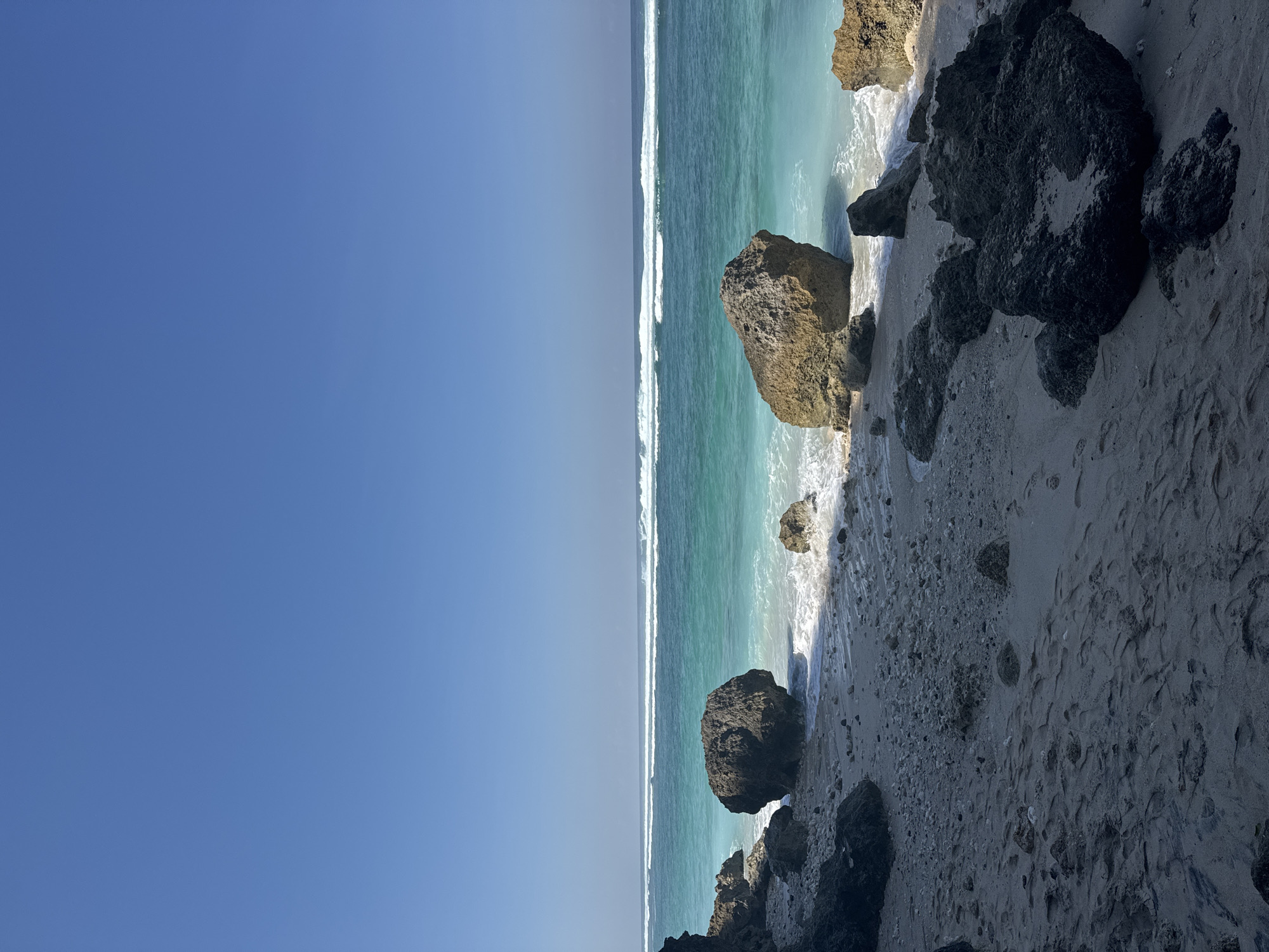 The image depicts a sunlit coastal landscape featuring a clear blue sky, a turquoise ocean with breaking waves, and a sandy, rocky beach. The upper third of the frame is dominated by a cloudless cerulean sky that gradually lightens towards the horizon. The middle third shows the ocean, characterized by its vibrant turquoise color near the shore and deeper blue further out. Multiple parallel lines of white-capped waves are visible, extending across the mid-distance of the water. Closer to the foreground, smaller white waves lap against the shore. The bottom third of the image is occupied by the beach, which consists of light-colored sand mixed with numerous dark grey and brownish rocks and lighter coral fragments. Footprints are visible on the sand in the lower central and left portions of the foreground. A prominent large, dark grey, rounded boulder is positioned in the mid-left at the waterline, partially submerged by a receding wave. Another large, lighter brown boulder sits in the mid-right, also at the water's edge. Darker, jagged rock formations are visible on the far left, suggesting a cliff or rocky outcrop, and in the lower right foreground. Shadows are cast across parts of the sandy beach and underneath rocks, indicating bright overhead sunlight. No individuals or animals are present in the scene. The location is an unidentifiable coastal area.
