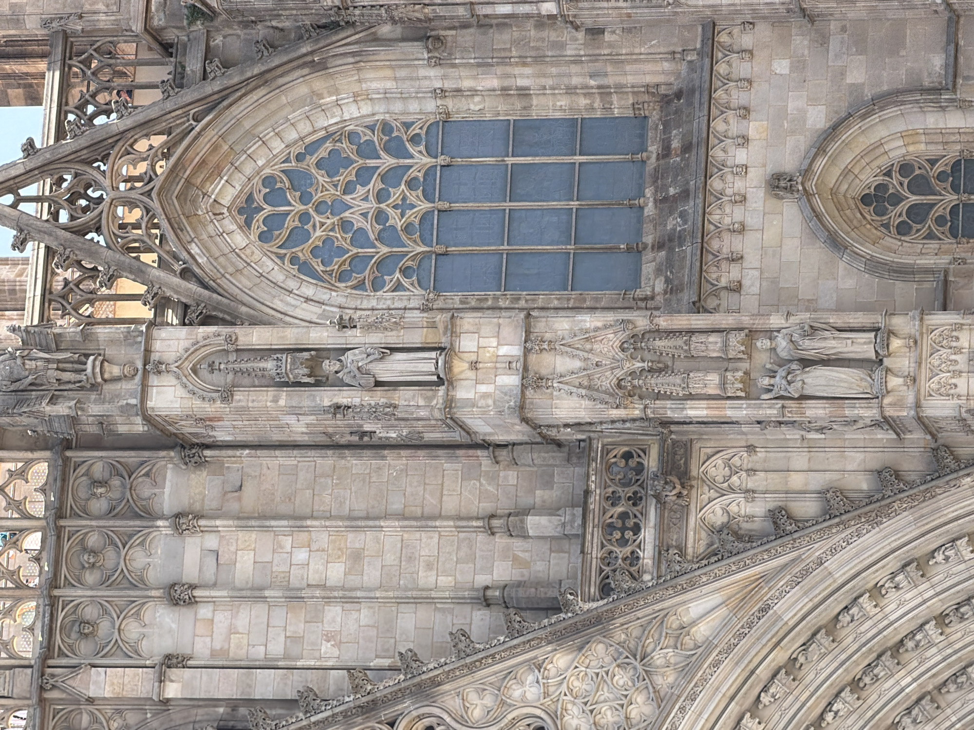 The image shows a detailed view of the exterior of the Barcelona Cathedral, located in the Gothic Quarter of Barcelona, Spain. The architecture is characterized by Gothic style, featuring intricate stonework, pointed arches, and decorative tracery.
A large, ornate Gothic window with blue glass is prominently visible on the right side of the image. The window's tracery forms a complex, symmetrical pattern. To the left of the window, there is a niche containing a statue of a robed figure, likely a saint or religious icon.
Further to the left, sections of the cathedral's facade reveal carved stone blocks forming columns and decorative elements. There are multiple niches containing statues of various figures, some appearing to be robed and holding objects. These statues are integrated into the architectural design, adding to the facade's intricate detail.
The lower left portion of the image shows a section of a large, curved archway, also adorned with detailed carvings and smaller statues within arched recesses. The overall impression is one of grand religious architecture with a high level of craftsmanship. The lighting suggests it is daytime, with shadows indicating the direction of the sun. No people are clearly identifiable as distinct individuals within the scene, though the statues represent human figures.
