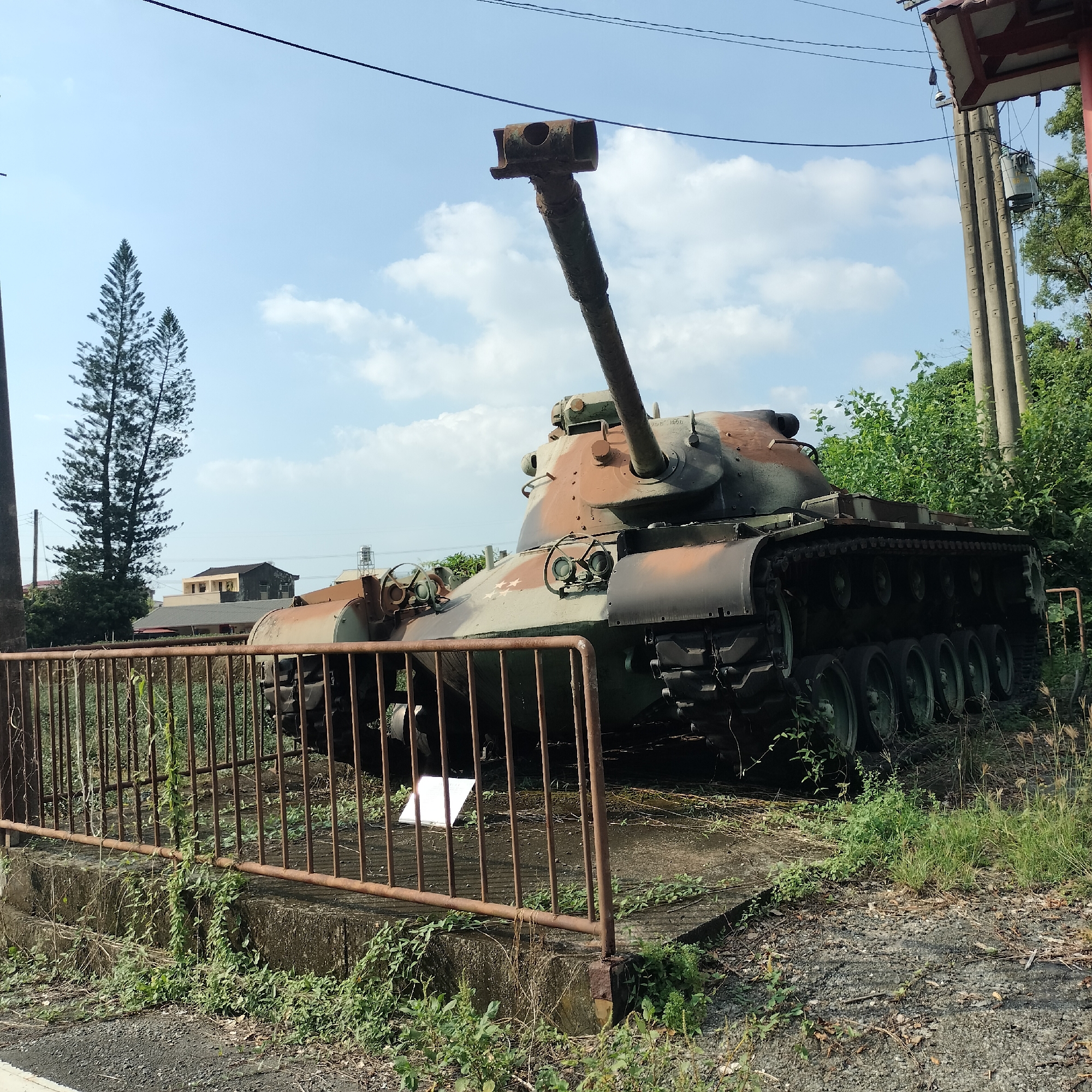 An abandoned military tank, camouflaged in brown and green, rests on a concrete platform behind a rusty metal fence. The tank is situated in an outdoor environment, with lush green foliage and trees surrounding it. In the background, buildings and power lines are visible under a clear blue sky with scattered clouds. The overall scene suggests a location in Zhushan Township, Taiwan.