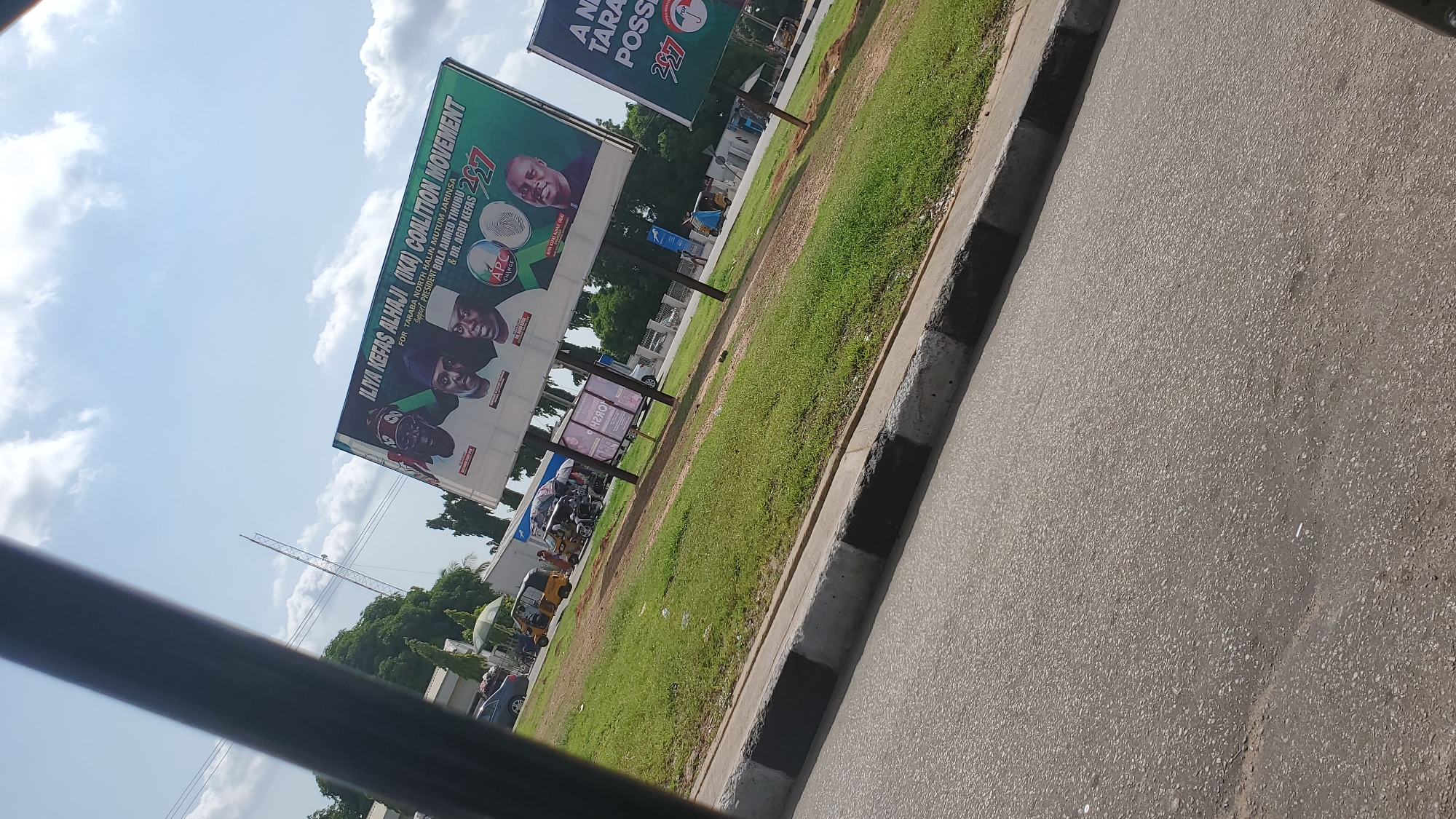 This media file shows a political billboard in Jalingo, Nigeria. The billboard, which is tilted and partially obscured by the foreground, features images of three men and text promoting "ILIYA KEFAS ALHAJI (IKA) COALITION MOVEMENT." It also includes slogans like "FOR TARABA NORTH HALIN MUTUM JARINSA," "Support PRESIDENT BOLA AHMED TINUBU," and "& DR. AGBU KEFAS." The logo for "APC" (All Progressives Congress) and the number "2027" are also visible.
To the right of the billboard, a paved road with a concrete curb is prominent. Beyond the road, a patch of green grass is visible, bordered by trees in the distance. Several vehicles, including what appear to be auto-rickshaws, are parked or moving in front of a building behind the grass. The sky is bright with scattered clouds, suggesting it is daytime. The overall scene depicts a public space in Nigeria, likely a street, with a strong emphasis on political campaigning.