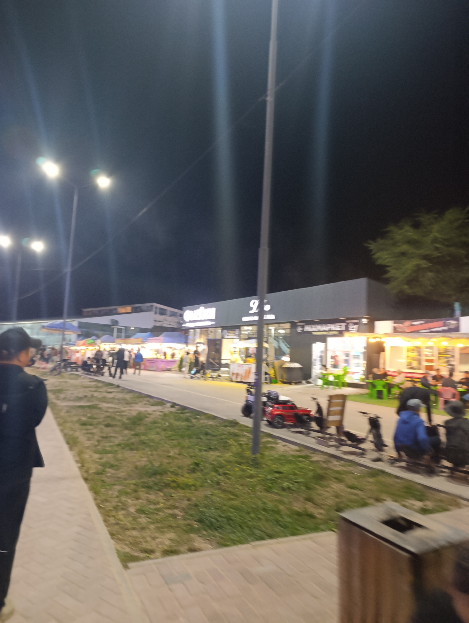 The image depicts a nighttime scene at a market or commercial area in Bosteri, Kyrgyzstan. Several food stalls and shops are visible, illuminated by artificial lights. People are milling about, some browsing the stalls, while others are seated at tables. Various vehicles, including a red ride-on toy car and scooters, are present in the foreground and along the pathway. The overall environment suggests a lively evening atmosphere at an outdoor gathering place.