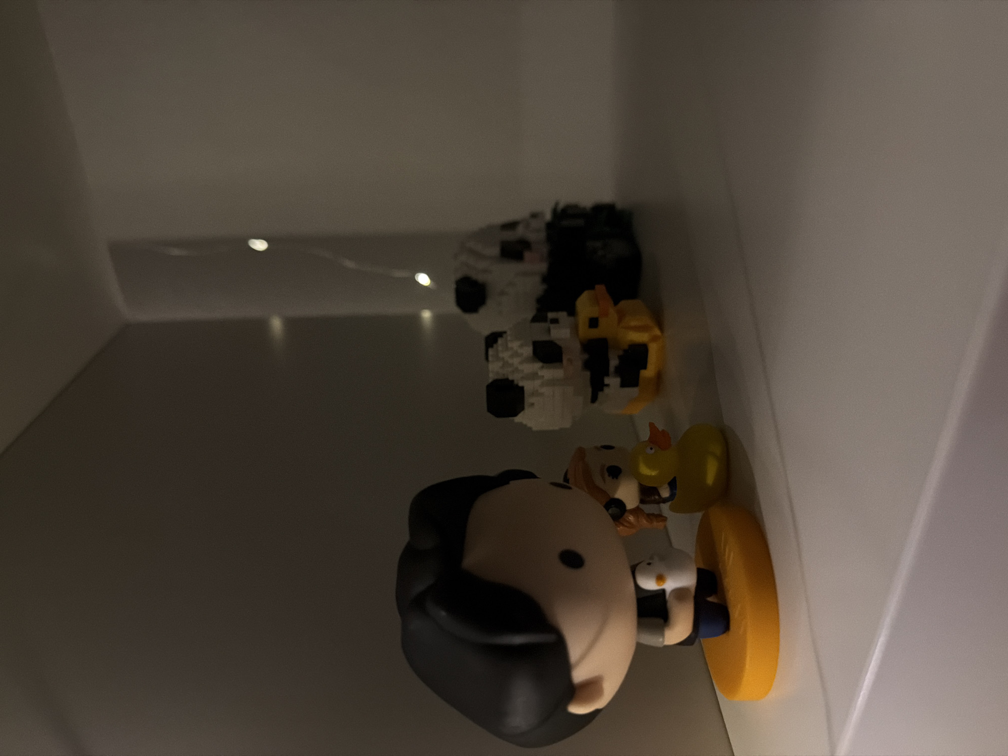 An indoor, eye-level medium shot captures a shelf with several figurines and a string of lights. On the left, a black-haired chibi-style figurine stands on an orange oval base, holding a white object. To its right is a smaller figurine with red hair. In front of the red-haired figurine is a yellow rubber duck with an orange beak and eyes. Further to the right are two pixelated panda figurines, one with a yellow element on its chest. A string of small, white lights is draped vertically on the right side of the frame, with some lights blurred in the background. The shelf is white, and the background is dark, suggesting a dim lighting environment. The scene is devoid of any indicators of its specific location within Kyiv, Ukraine.