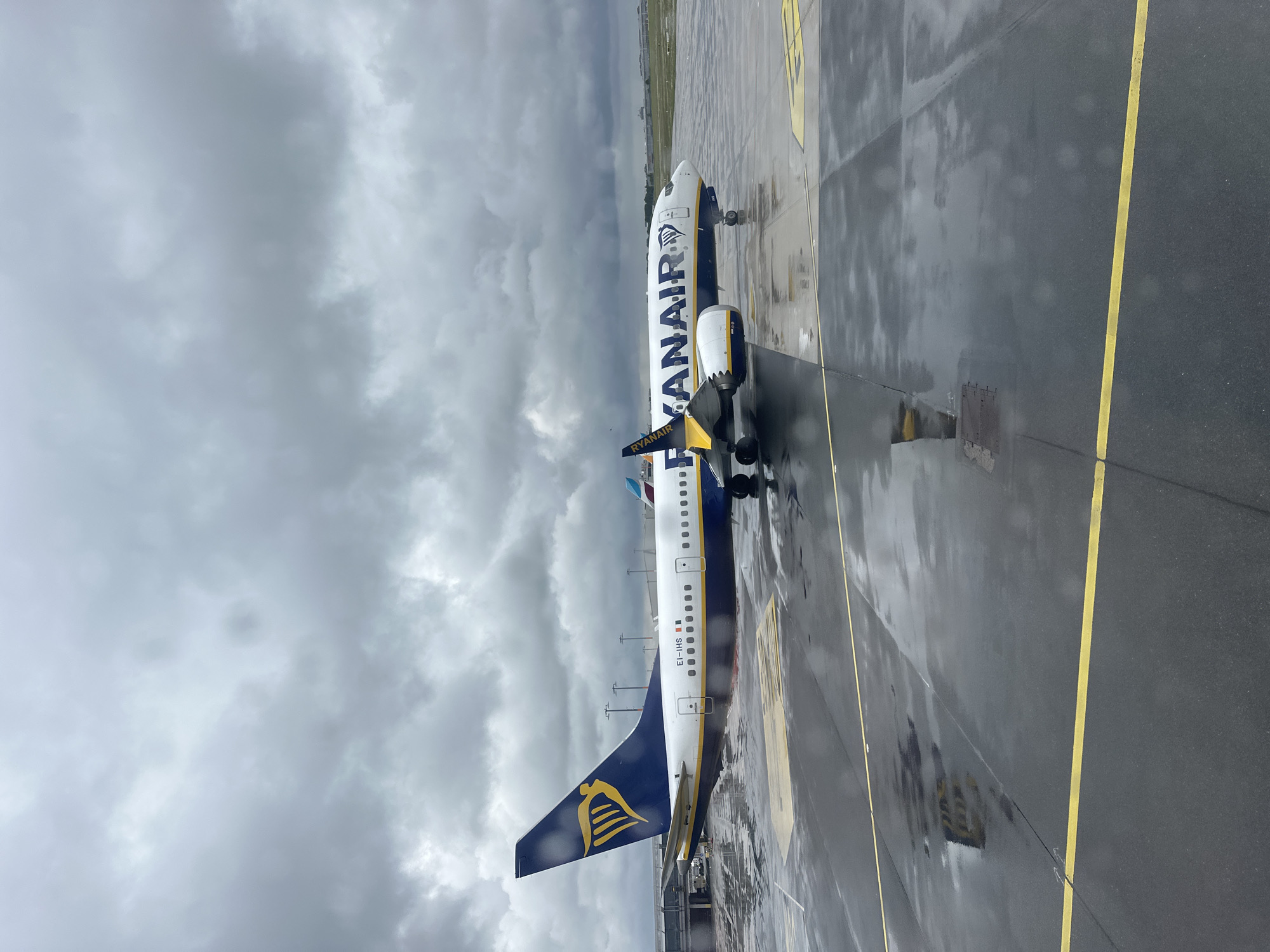 This image shows a Ryanair airplane parked on a wet tarmac at Hamburg Airport (HAM), Germany. The sky is heavily overcast with dark, dramatic clouds, suggesting recent or impending rain. The tarmac itself is reflective, with puddles of water and yellow painted markings, indicating an active airport environment. The airplane is the primary subject, its blue and yellow livery clearly visible, along with the Ryanair logo on the tail fin. The registration number "EI-IHS" is visible on the fuselage. Other airport infrastructure, such as lights and a jet bridge, can be seen in the background, along with a glimpse of a second airplane. The overall scene conveys a sense of a typical, slightly dreary day at a busy European airport. There are no people visible in the image.