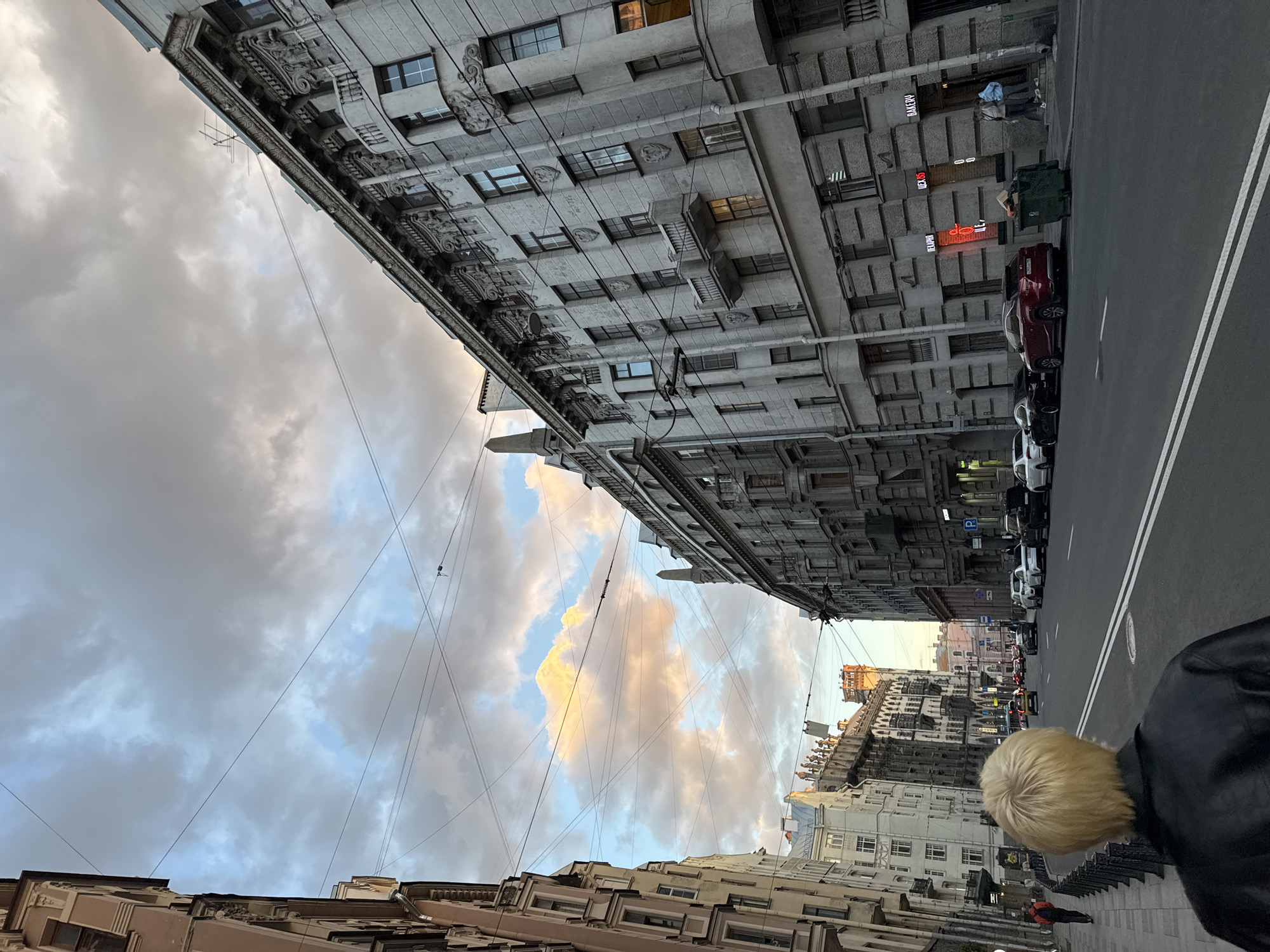 The image shows a street scene in Saint Petersburg, Russia, on a cloudy day with patches of sunlight illuminating the sky. The perspective is from behind a person with blonde hair, facing down a wide street lined with ornate, multi-story buildings. The architecture of the buildings features detailed facades with balconies, decorative moldings, and numerous windows. Overhead, a network of electrical wires crisscrosses the sky.
In the foreground, the person in the black jacket is walking on the sidewalk. The street itself is paved and has double white lines marking the lanes. Several cars are parked along the right side of the street and some are driving in the distance. The buildings on the right have storefronts with illuminated signage, one reading "BAKERY." On the left side of the street, more buildings with similar architectural styles are visible. In the background, the street extends into the distance, with more cars and buildings. The sky is filled with dramatic clouds, with some areas tinged with warm colors from the setting or rising sun. The overall impression is of a European city street, characteristic of Saint Petersburg's historical urban landscape.