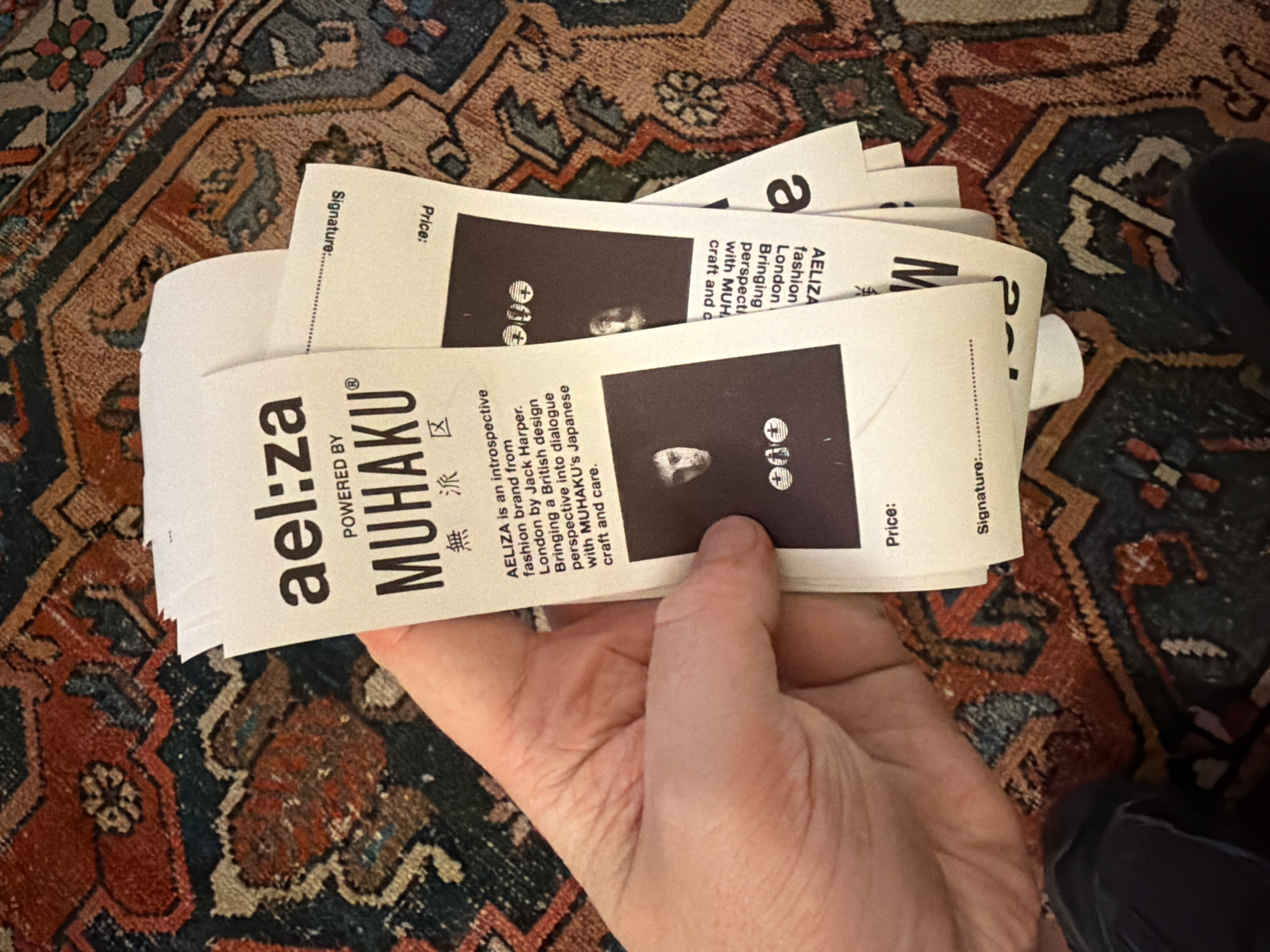 Here's a description of the image:
A hand holds a stack of white tags or labels printed with text and a dark square containing what looks like a face with a plus sign over the eyes. The text reads "ael:za POWERED BY MUHAKU," with Japanese characters and "AELIZA is an introspective fashion brand from London by Jack Harper. Bringing a British design perspective into dialogue with MUHAKU's Japanese craft and care." The reverse side has "Price:" and "Signature:" prompts. The background is a detailed oriental rug. The environment gives no indication that the image was taken in the city of New York, United States of America.