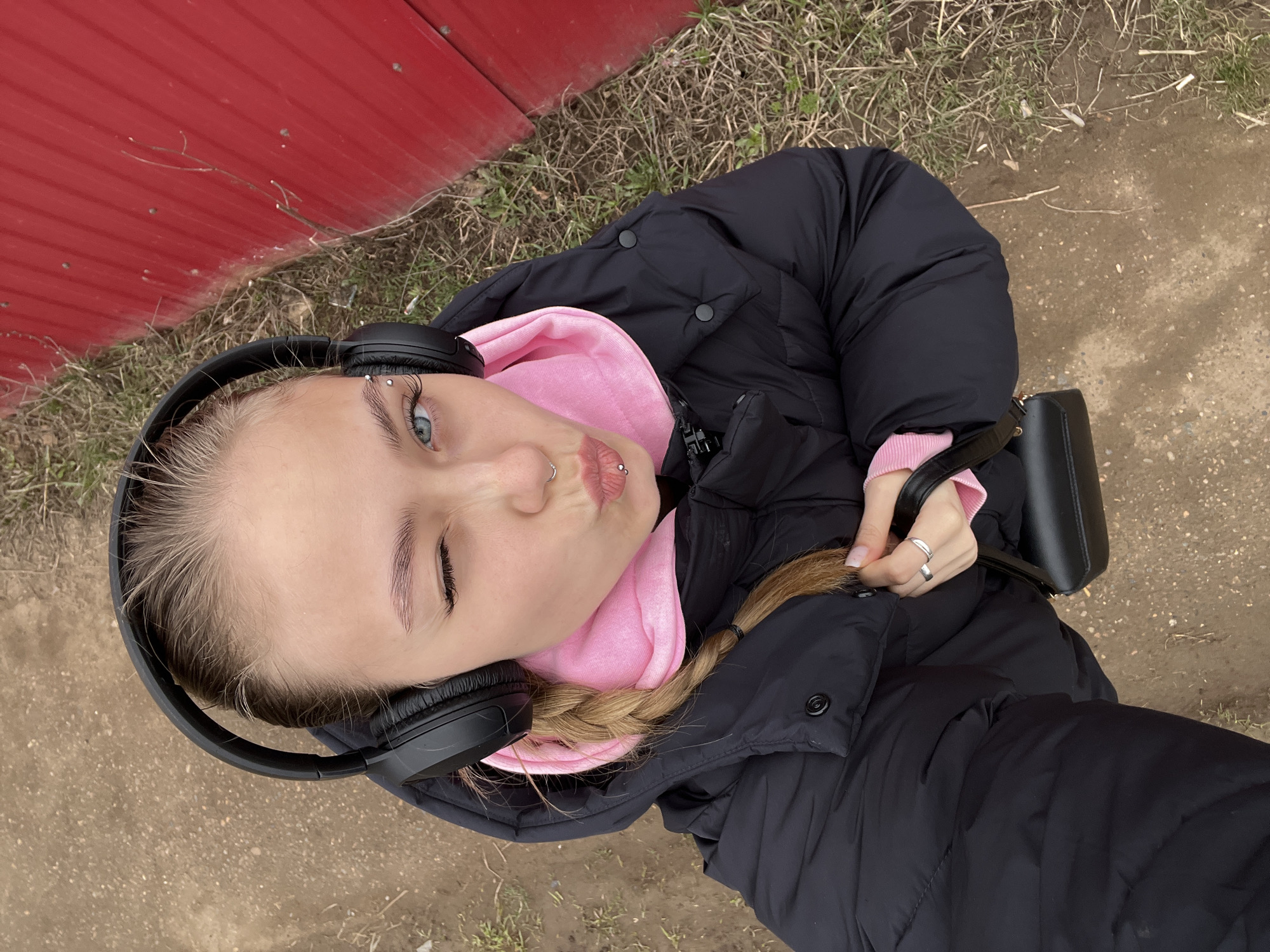 A young woman is seen from a high-angle perspective, likely taking a selfie outdoors. She has blonde hair, styled in a braid, and blue eyes. She is wearing a black puffer jacket over a pink hoodie, along with black headphones. She has piercings in her eyebrow, nostril, and lip. Her right eye is winking. She is standing on what appears to be a dirt or gravel path. A red wall is visible on the left edge of the frame. The location could be Фоки, Russia.