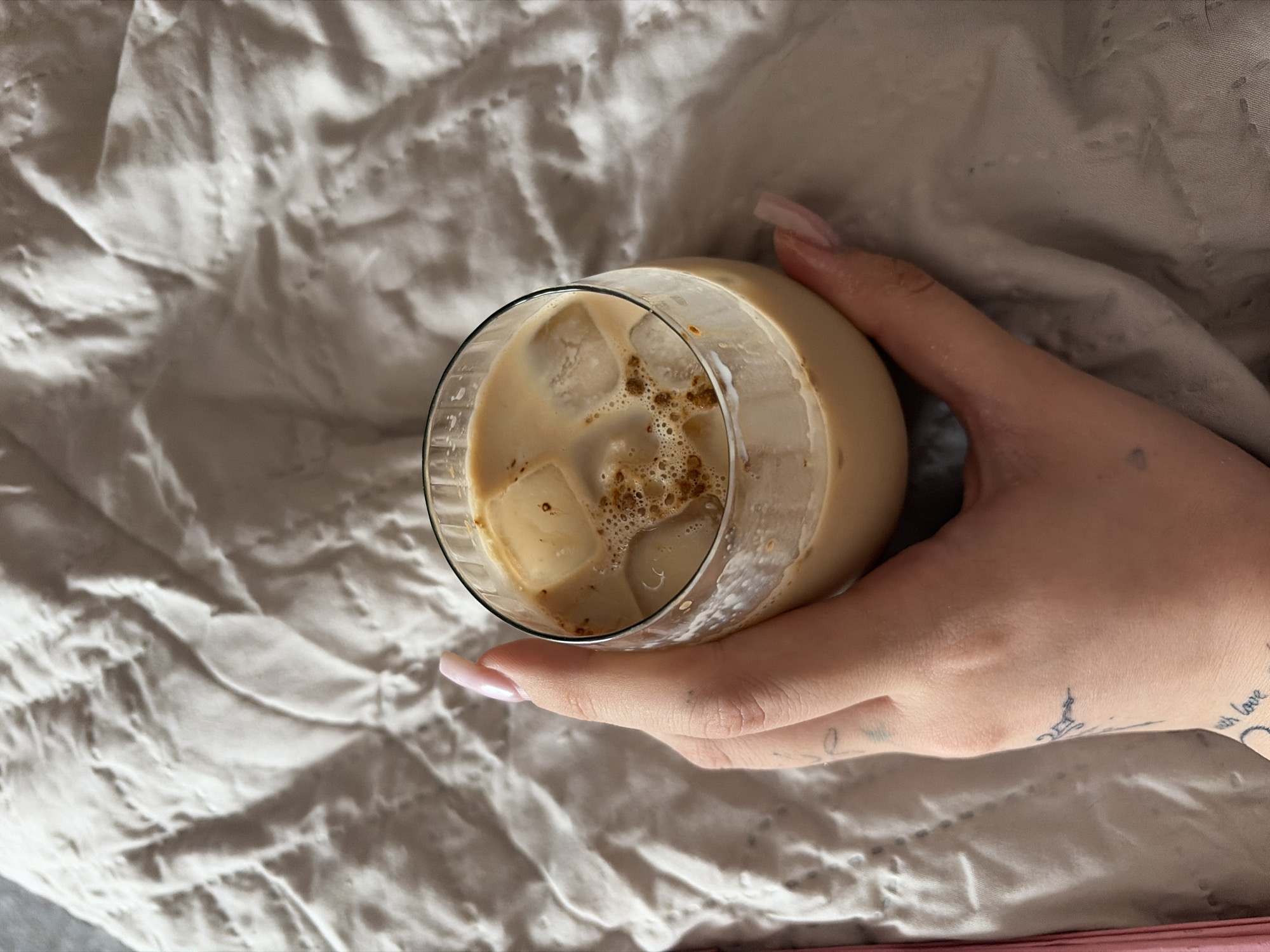 The image displays an overhead, close-up view of a person's hand holding a glass of iced beverage, set against a rumpled fabric background. The hand, likely a right hand, is positioned towards the bottom right of the frame, gently gripping a clear glass. The person has long, almond-shaped fingernails painted in a light, neutral color, possibly pale pink or nude. Several small tattoos are visible on the hand and wrist; one appears to be a silhouette, possibly a building like the Eiffel Tower, on the back of the hand near the index finger, and a partial text tattoo reading "with love" is visible on the wrist.
The glass contains a milky, light-brown drink, indicative of an iced coffee, latte, or similar beverage. Several ice cubes are visible within the drink, and the surface features small, dark brown specks or froth, likely from coffee.
The background is composed of a light-colored, possibly off-white or light grey, rumpled fabric, suggesting a bedsheet, blanket, or soft furnishing. The folds and shadows on the fabric create a soft, textured surface.
The activity depicted is a person holding and presumably enjoying a cold drink, suggesting a moment of relaxation or leisure in an indoor setting. No specific time of day or weather conditions can be determined from the lighting. The overall mood is one of comfort and calm. The location context for this scene is Chaikovsky, Russia.