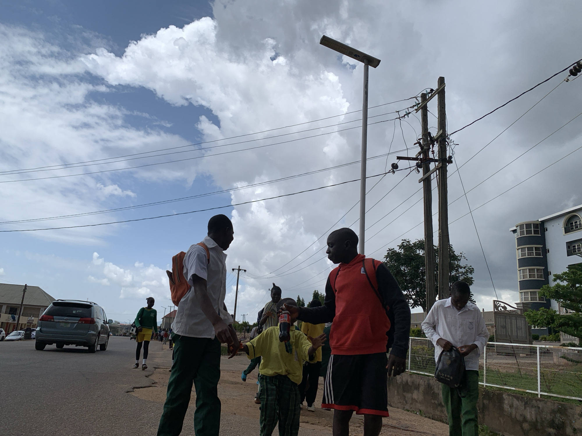 The image captures a street scene in Jos, Nigeria, under a cloudy sky. Several individuals, appearing to be students in school uniforms, are present. In the foreground, two young men are interacting. One, wearing a white shirt and green trousers, has an orange backpack slung over his shoulder. The other, in a red hoodie and black shorts with white stripes, is facing him. Between them, a younger child in a yellow shirt and checkered trousers is holding a bottle of soda.
Further in the background, other students are visible walking on the street and sidewalk. A grey minivan is driving on the road, and residential buildings and a modern, multi-story building with rounded balconies are present in the background. Electrical poles and overhead wires crisscross the sky, indicating urban infrastructure.
The time of day appears to be afternoon, given the angle of the light and the presence of shadows. The weather is overcast, with dramatic clouds suggesting the possibility of rain. The overall atmosphere seems casual, with the students engaged in interaction as they move along the street. There is no visible text in the image.