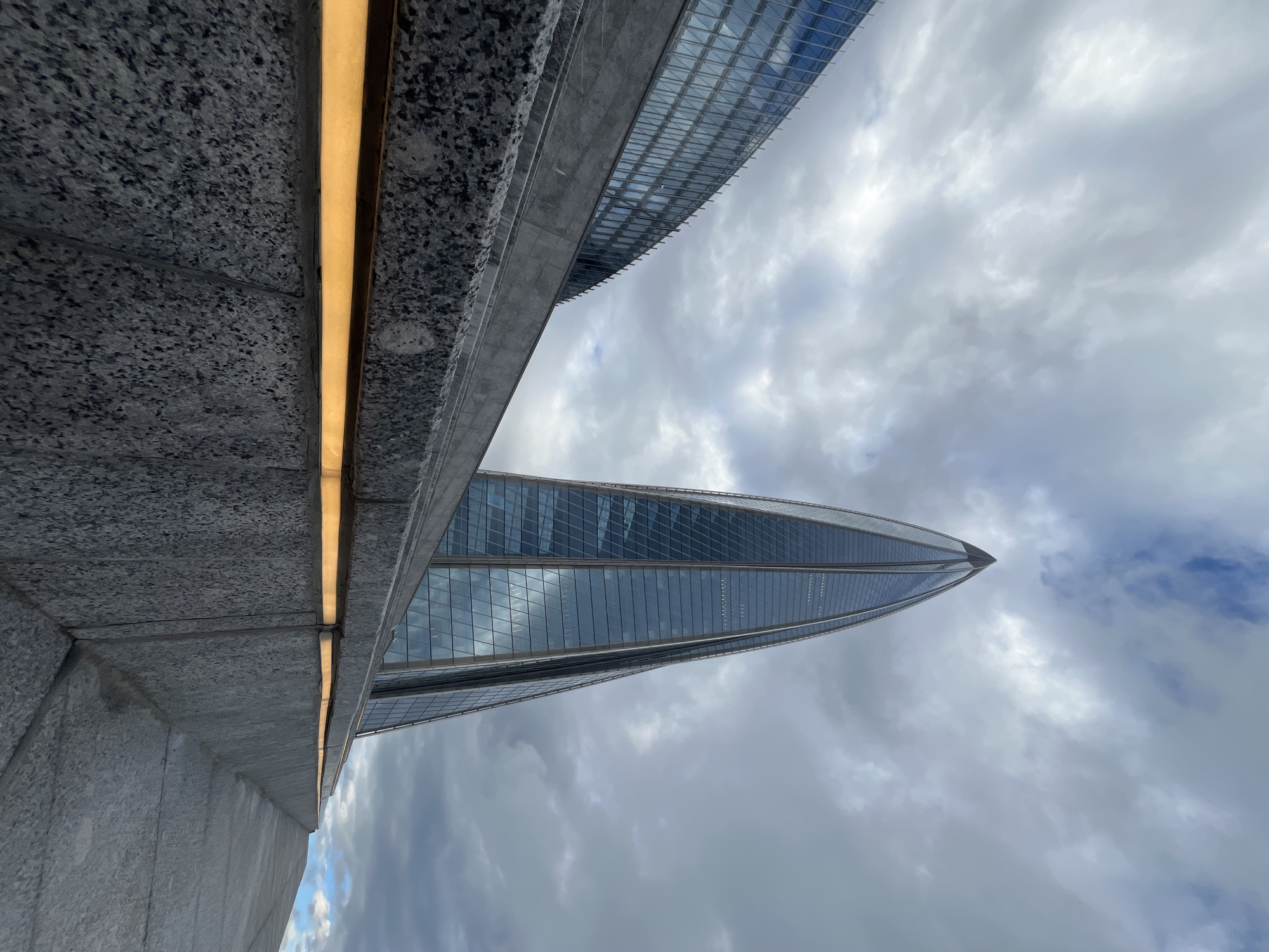 The image shows a low-angle view of the Lakhta Center skyscraper in Saint Petersburg, Russia, against a cloudy sky. The skyscraper, a prominent architectural landmark, is a tall, glass-clad building with a distinctive pointed top that tapers towards the sky. In the foreground, a large, grey granite structure with integrated warm-toned lighting runs along the bottom of the frame, creating a contrast with the modern skyscraper. The granite structure appears to be part of a plaza or walkway leading towards the building. Another building with a reflective glass facade is visible to the left of the skyscraper. The overall scene captures a modern urban landscape with a focus on architectural grandeur and the interplay of light and shadow.