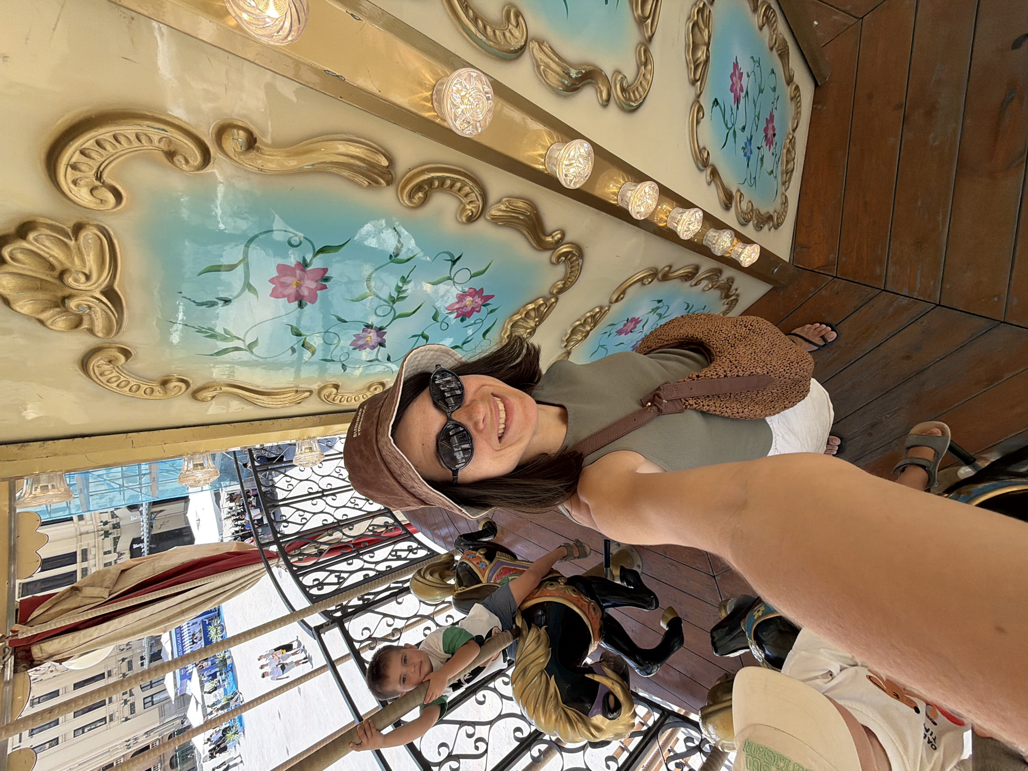 This is a selfie taken by a woman on a carousel in Valencia, Spain. She is wearing a brown hat and sunglasses, and a green shirt. A young boy, wearing a white and green t-shirt, is riding on a carousel horse next to her. The carousel is decorated with ornate gold trim and painted floral designs. The woman appears happy and excited, smiling at the camera. The image is taken during the daytime, and the weather appears to be sunny. The background shows buildings and a bright sky, suggesting an outdoor location. There is no visible text in the image.
