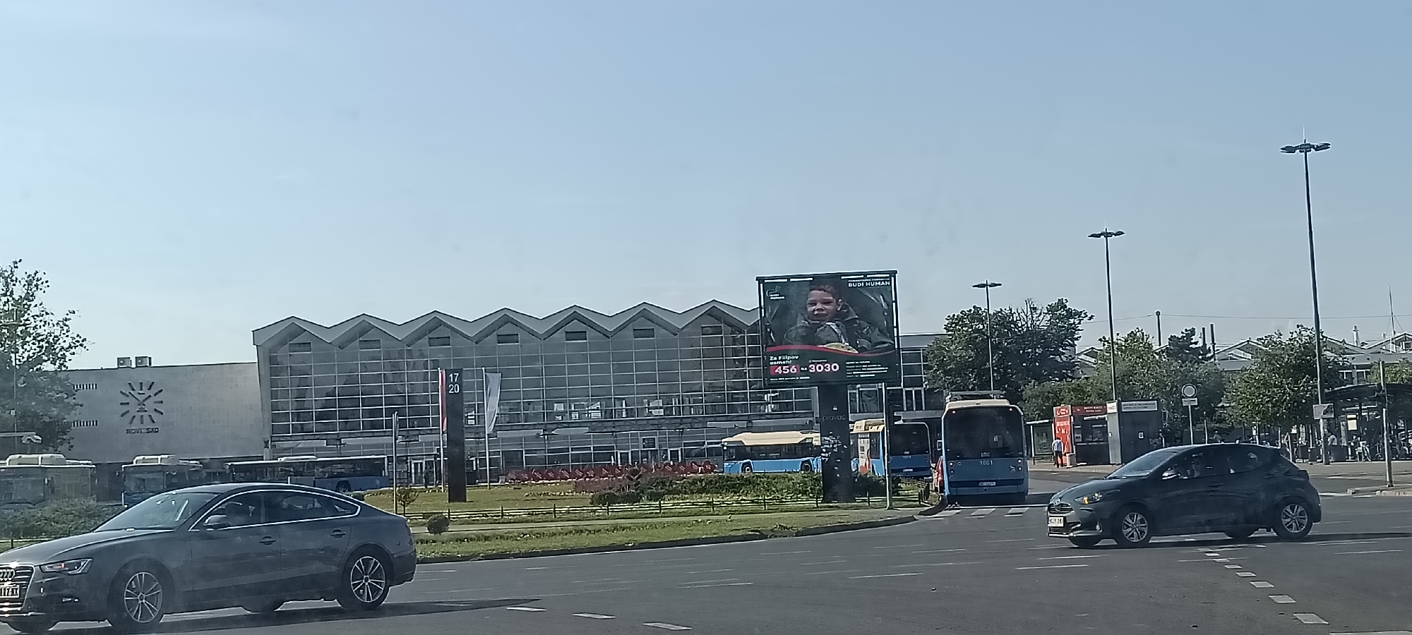 The image depicts a street scene in Novi Sad, Serbia, near the bus station. A large modern building with a distinctive zigzag roofline and a glass facade dominates the background. To the left of this building, a large clock face is visible on a concrete wall, with "NOVI SAD" written below it. In front of the buildings, several blue buses are parked or driving, indicating a transportation hub. A prominent billboard displays an advertisement featuring a young boy, with text including "BUDI HUMAN" and a phone number. In the foreground, two cars are on the road: a dark gray Audi sedan on the left and a dark gray hatchback on the right. The sky is clear and blue, suggesting a sunny day. The overall environment appears to be urban, with streetlights and trees also visible.