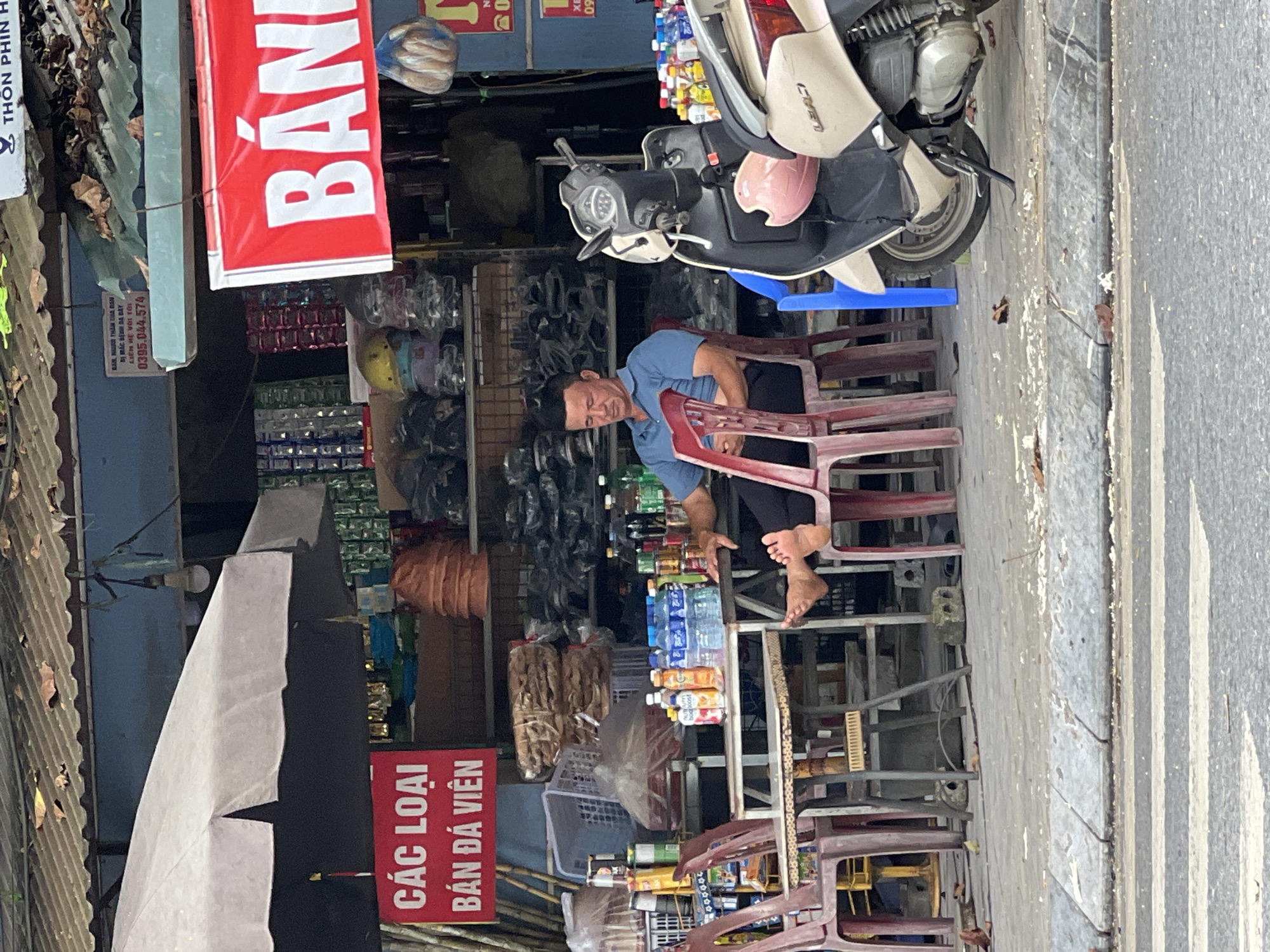 A middle-aged Vietnamese man with short, dark hair is sitting on a plastic chair at a table outside a shop in Tân Quang Commune, Vietnam. He is wearing a blue polo shirt and dark pants, with his bare feet resting on the seat of another chair. The shop appears to be selling various goods, with shelves of products visible in the background. A red sign with white text that reads "CÁC LOẠI BÁN ĐÁ VIÊN" (meaning "Types of ice cubes for sale") is prominently displayed. A red and white banner with the word "BÁ" visible is also hanging from the shop. A light-colored scooter is parked to the right of the man, with a pink helmet resting on its front. The scene is outdoors, with a sidewalk in the foreground and a street with a crosswalk visible. The shop has a corrugated metal roof.
