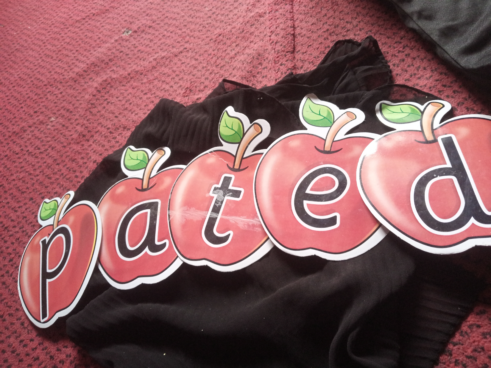 A close-up, eye-level shot captures a portion of the word "pated" spelled out in individual apple-shaped cutouts. Each cutout is red with a white border and features a green leaf and a brown stem. The letters are black and stylized. From left to right, the visible letters are "p", "a", "t", "e", and "d". The cutouts are arranged diagonally across a dark-colored, ribbed fabric, likely clothing. The background is a textured, reddish surface, possibly a carpet or rug. The lighting is neutral, and there are no visible people or indications of the specific location in Jalingo, Nigeria.
