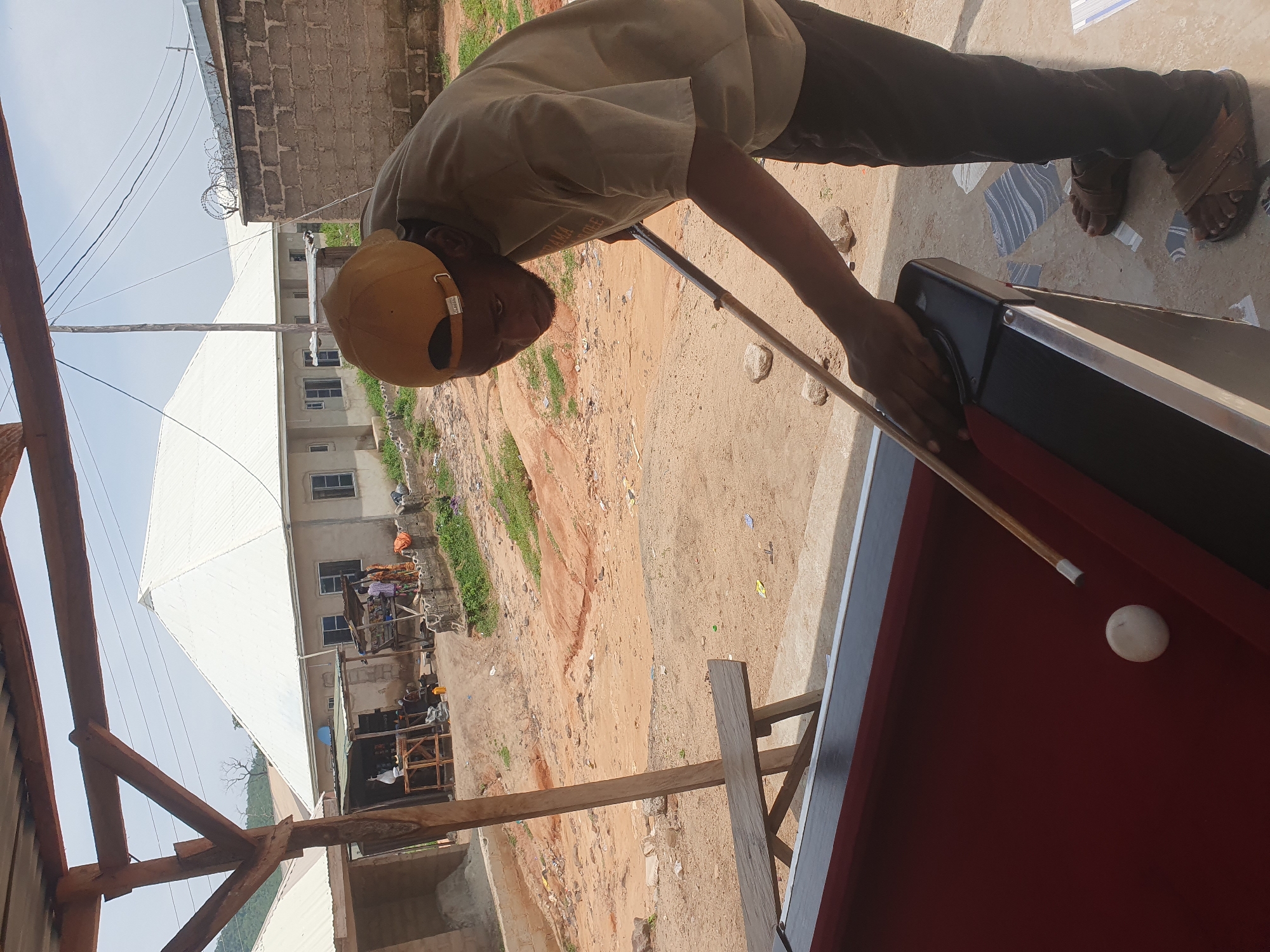A man wearing a brown t-shirt and a tan baseball cap is playing pool outdoors in Lokoja, Nigeria. He is holding a pool cue and aiming at a white ball on a red felt pool table. The background shows a dusty street with several buildings and trees. The sky is clear and blue.