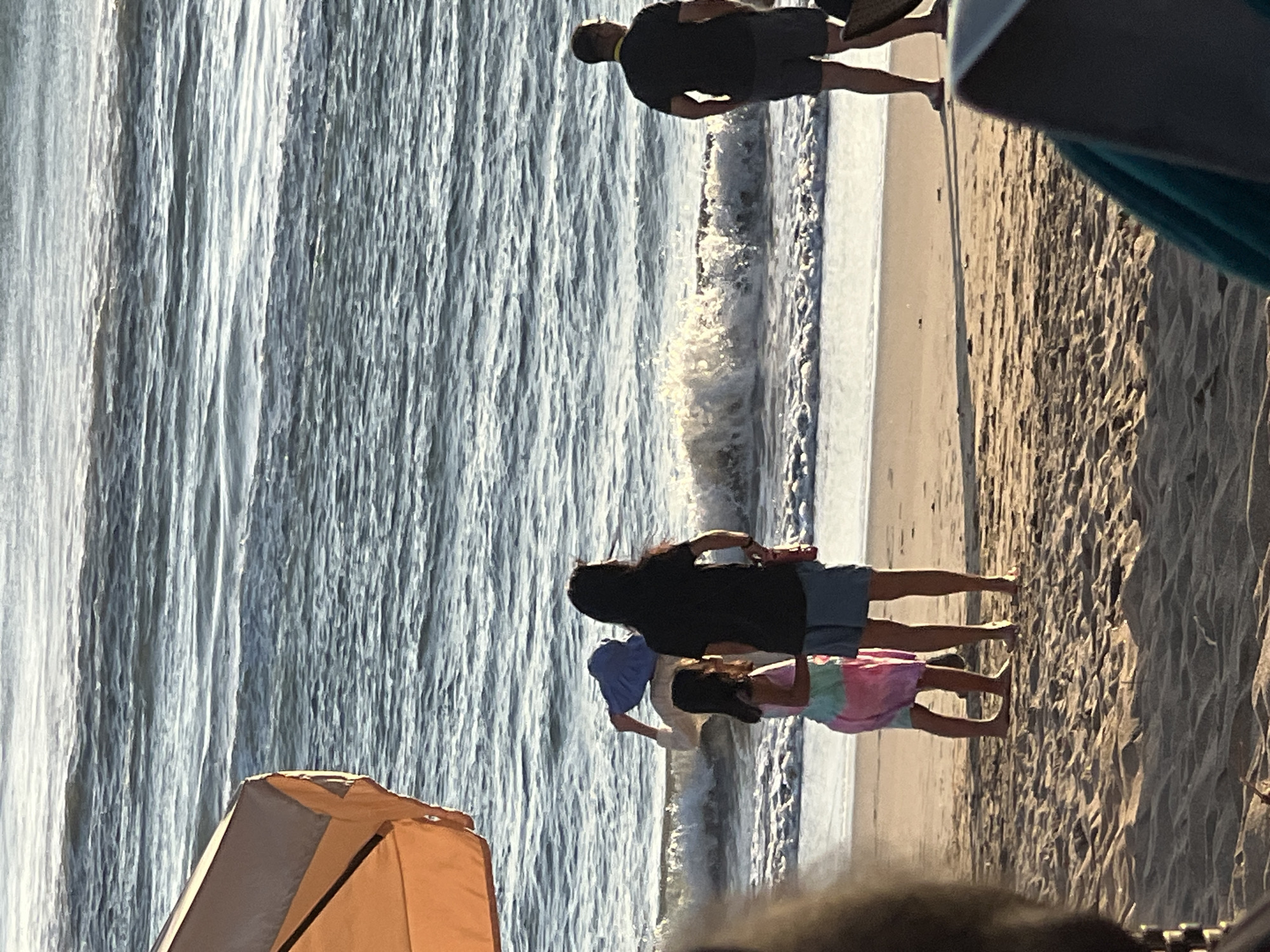 A group of four people are on a sandy beach in Kuta, Indonesia, with the ocean in the background. The water is choppy with waves breaking on the shore. In the foreground, a person's head and shoulders are visible, out of focus. To the right of the foreground, a beach chair with a blue fabric is visible.
In the middle ground, three people are standing on the sand near the water's edge. A woman with long dark hair and a black t-shirt and blue shorts is facing away from the camera. To her left, a young girl in a tie-dye pink, green, and blue dress is also facing away from the camera. Between the woman and the girl, a third person, whose face is not visible, is wearing a light-colored top and holding a blue hat over their head.
Further to the right in the middle ground, a man wearing a black t-shirt and grey shorts is standing and looking out at the ocean. He has glasses on and a yellow lanyard around his neck.
An orange beach umbrella is visible in the upper left corner of the image, casting a shadow on the sand. The sand is a light brown color and shows footprints and ripples from the wind. The lighting suggests it is late afternoon or early morning.