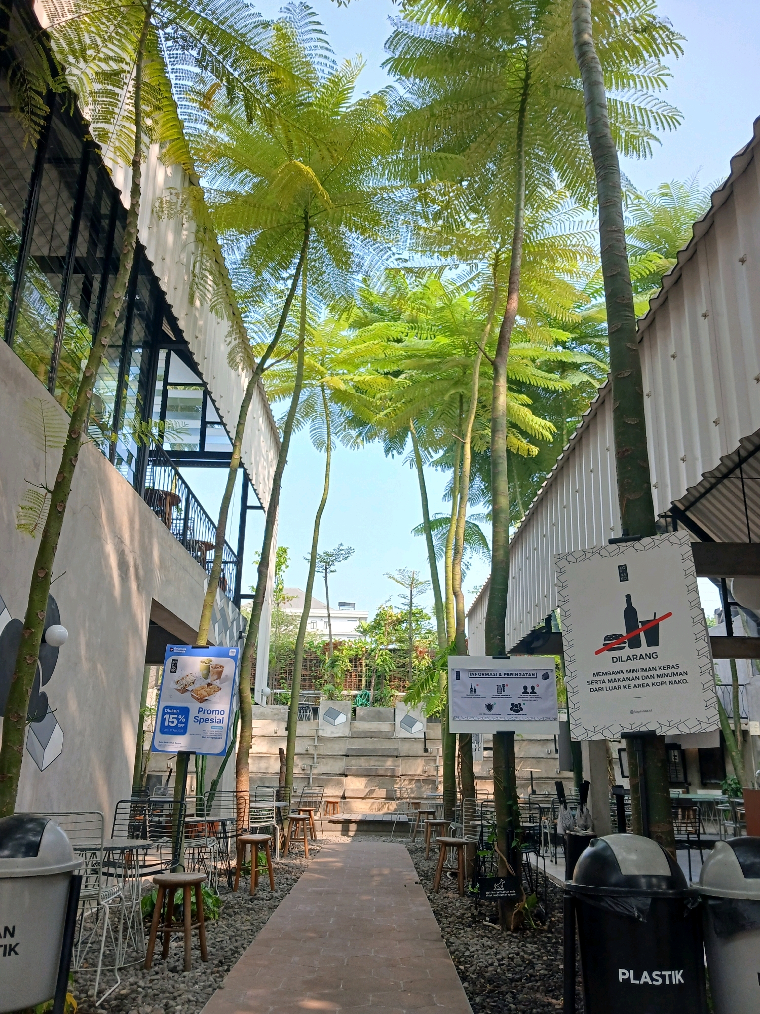 In Surakarta, Indonesia, a row of tables and chairs is set up outdoors at a coffee shop with a contemporary, industrial design. Tall, slender trees with feathery fronds create a canopy over the seating area. The architecture features corrugated metal siding and large glass windows. Two prominent signs are visible: one announces a "Special Promotion 15% OFF" with images of food items, and the other is a warning sign in Indonesian that reads "DILARANG MEMBAWA MINUMAN KERAS SERTA MAKANAN DAN MINUMAN DARI LUAR KE AREA KOPI NAKO" which translates to "FORBIDDEN TO BRING ALCOHOLIC DRINKS AS WELL AS FOOD AND DRINKS FROM OUTSIDE TO THE KOPI NAKO AREA". Black trash cans, one labeled "PLASTIK", are placed near the entrance. The ground is covered with gravel, and a paved walkway leads through the space. The sky is clear and blue. No people are present in the immediate vicinity of the tables and chairs.
