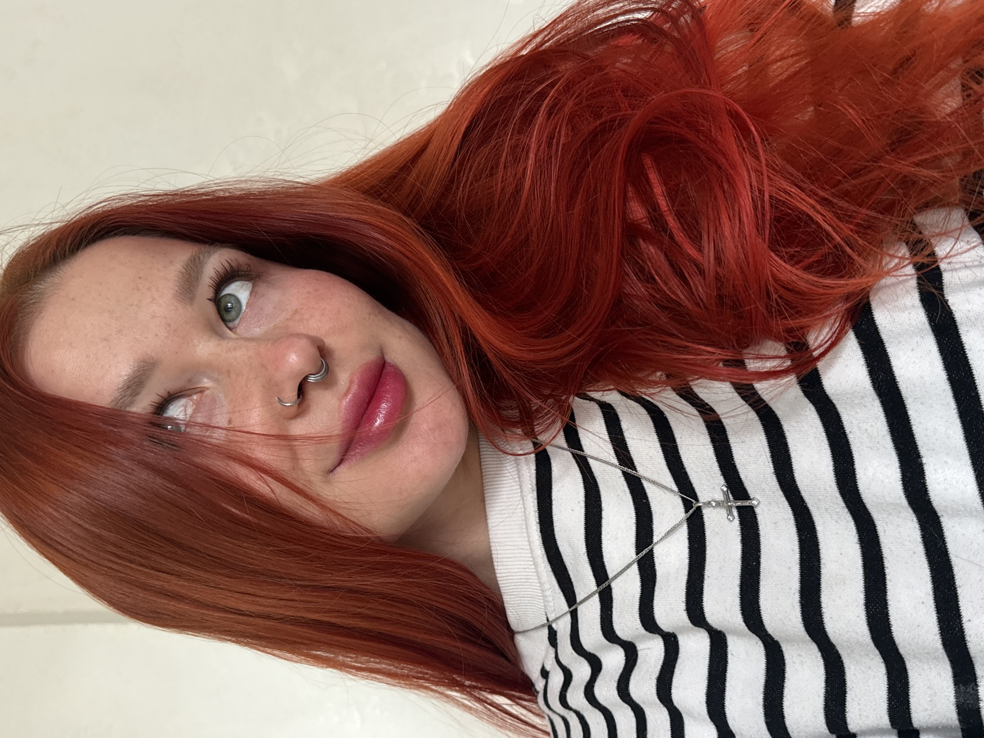 A young woman with fair skin, freckles, and vibrant red hair poses for a selfie. She wears a black and white striped shirt and a delicate silver necklace with a cross pendant. A silver nose ring is visible in her left nostril. Her lips appear glossy with a pink hue. Her gaze is directed slightly upward and to the left. The background is a plain white wall. There is no information in the image that indicates the location is Rostov-on-Don, Russia.