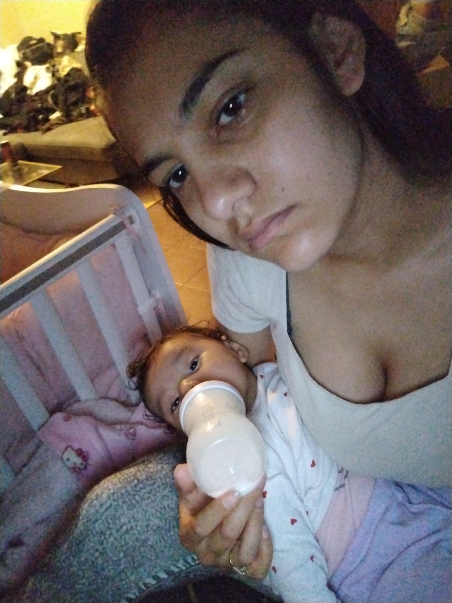 A woman takes a selfie with a baby who is drinking from a bottle. The baby is lying in a crib with a blanket featuring a Hello Kitty design. The woman is wearing a white top, and the baby is wearing a white outfit with small red hearts. The location is a domestic indoor environment. The city and state are unknown.