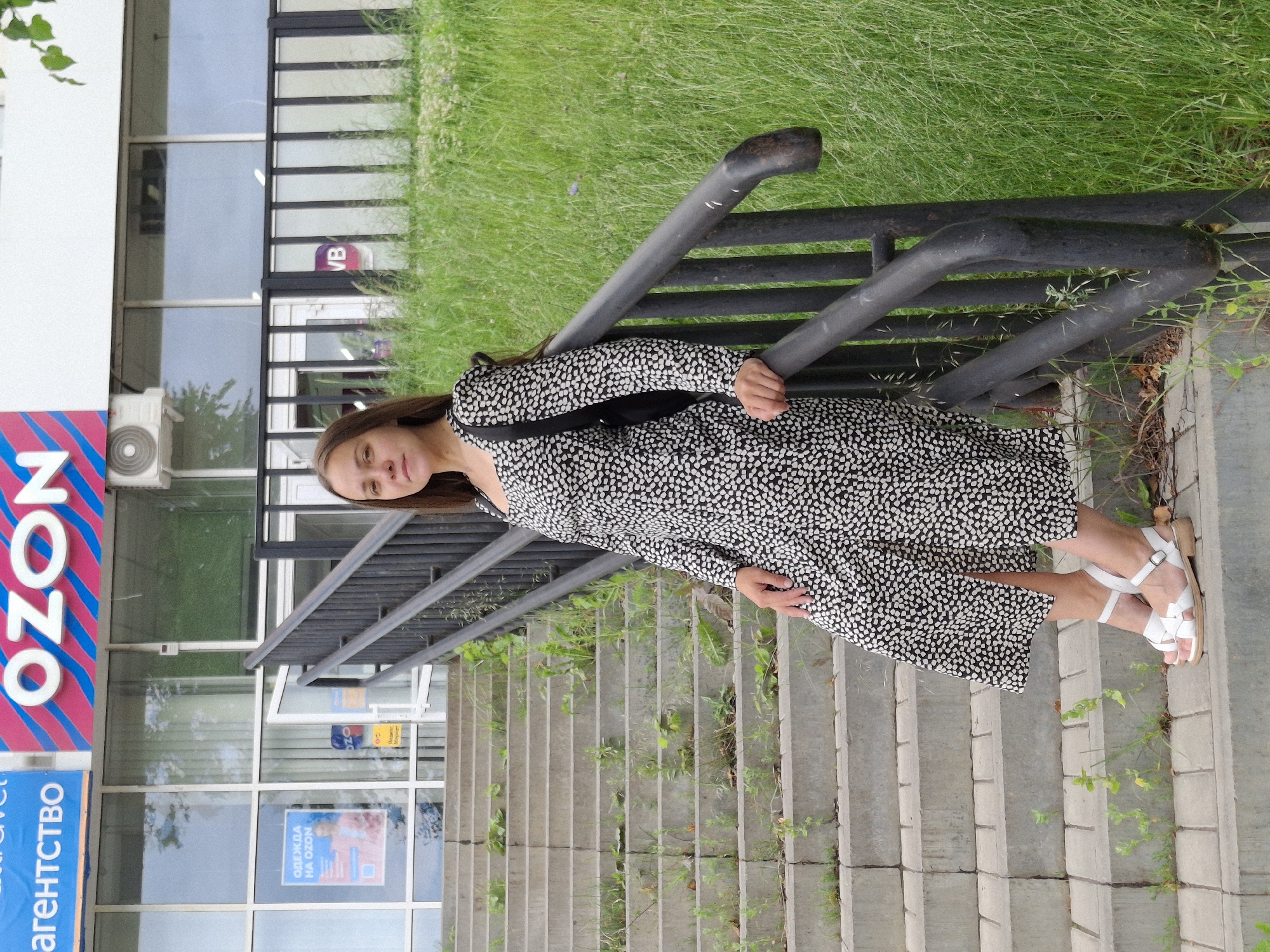 A young woman with long brown hair is standing on a concrete staircase outdoors, leaning against a black metal railing. She is wearing a black and white patterned dress and white sandals. The staircase leads up to a building with large windows, and signage with the word "OZON" is visible. The building appears to be a commercial establishment. The area surrounding the staircase is overgrown with green grass and some sparse vegetation. In the background, a street and more buildings can be seen. The overall impression is of a casual outdoor scene in an urban or suburban setting. The location is identified as Chaikovsky, Russia.