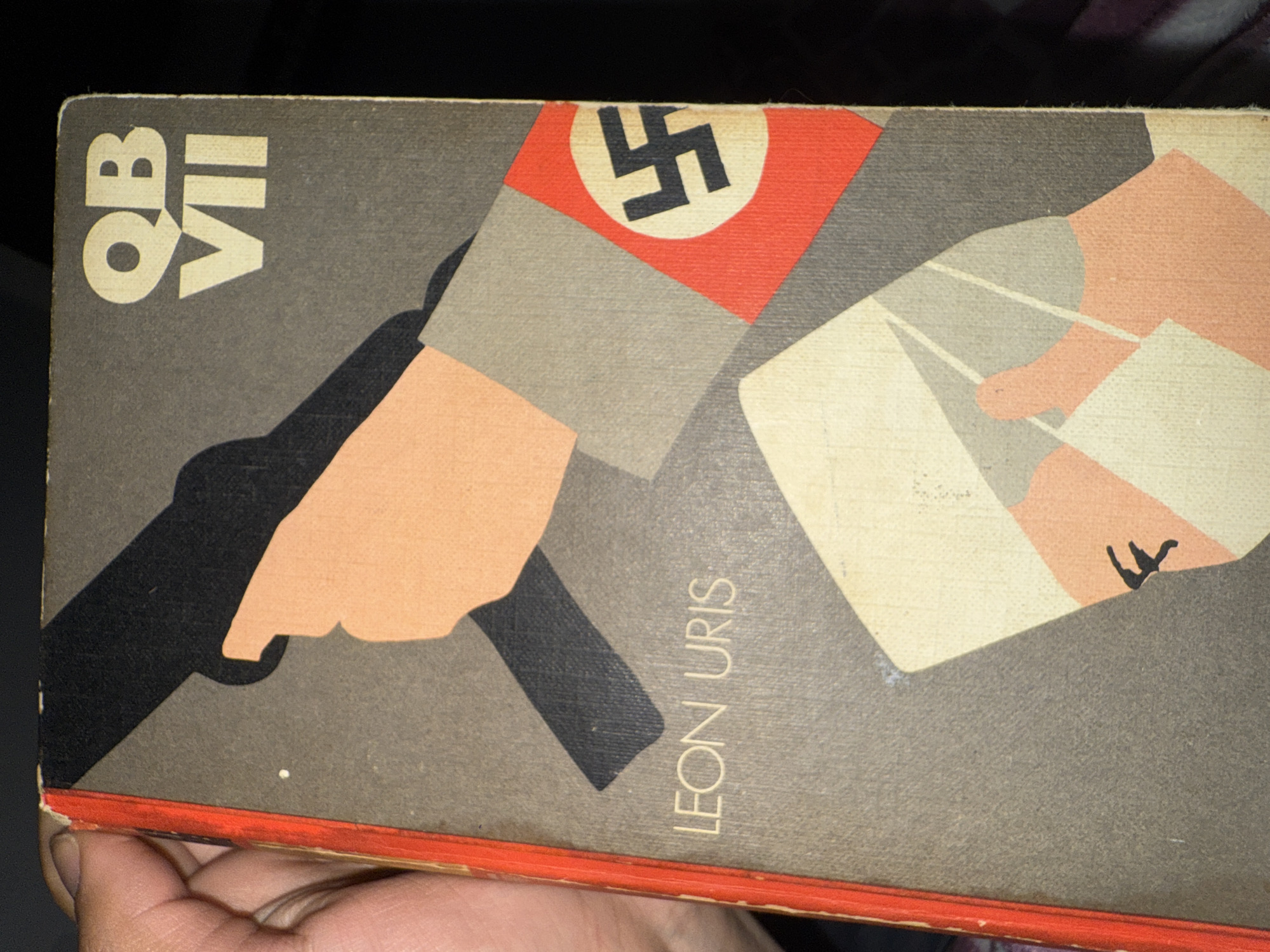 The image shows the cover of the book "QB VII" by Leon Uris. The cover art features a stylized depiction of a person holding a pistol, with a swastika on their arm. The book's title, "QB VII," is printed in bold lettering on the left side of the cover, and the author's name, "Leon Uris," is printed vertically on the right side. The background of the cover is a mottled grey color.
The location in the city of Barcelona, Spain is not discernible from the provided image.
