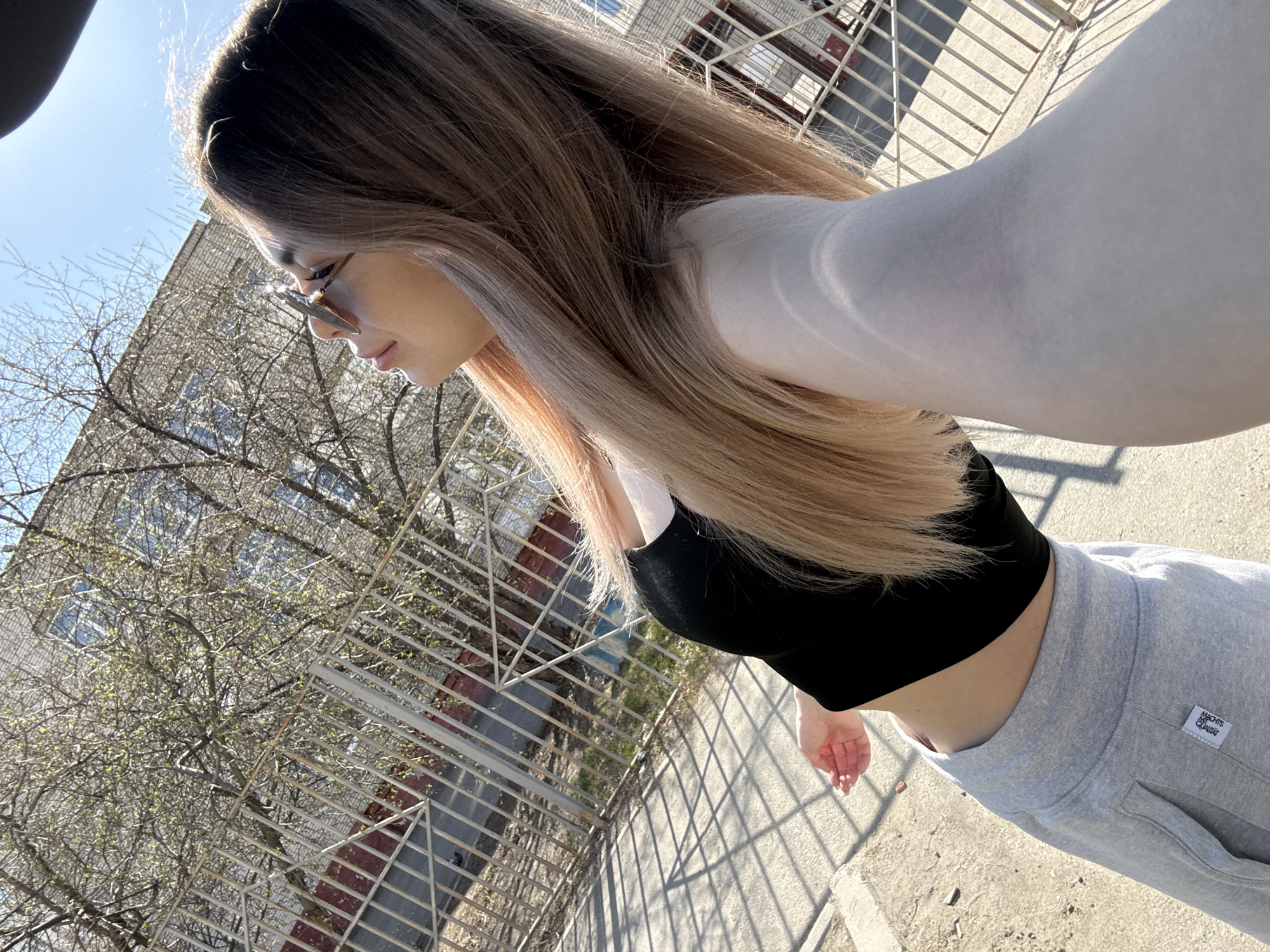 A woman is standing outside near a building with several windows. She has long brown and blonde hair and is wearing sunglasses. She is wearing a black top and grey sweatpants. The area is partially shaded by a bare tree and a metal fence.